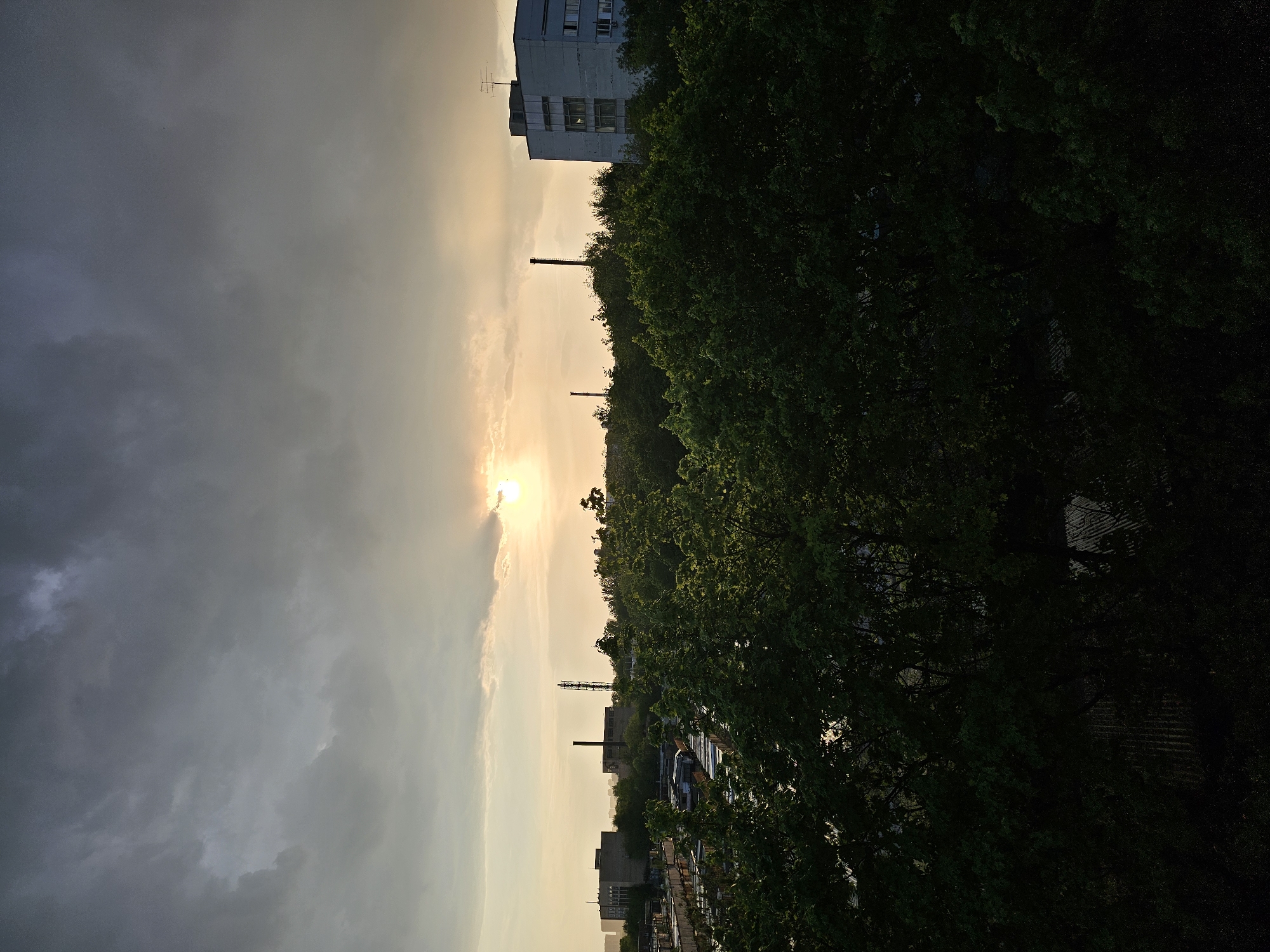 The image captures a view in Khimki, Russia, at what appears to be sunset or sunrise. The scene includes industrial elements juxtaposed with natural features.
The sky is divided, with dark, cloudy conditions dominating the left side of the image and a brighter, sunlit area visible towards the center. The sun is partially obscured by clouds, creating a bright glare.
The right side of the image is characterized by lush, green trees that fill a significant portion of the frame. Several industrial chimneys or towers punctuate the skyline, interspersed among the trees. In the mid-ground, rooftops of buildings and a partial view of vehicles are visible. Further back, a taller, light-colored building with visible windows and what appears to be an antenna on top stands out against the sky.
The photo is taken from an elevated perspective, overlooking the scene. The orientation of the image is vertical.