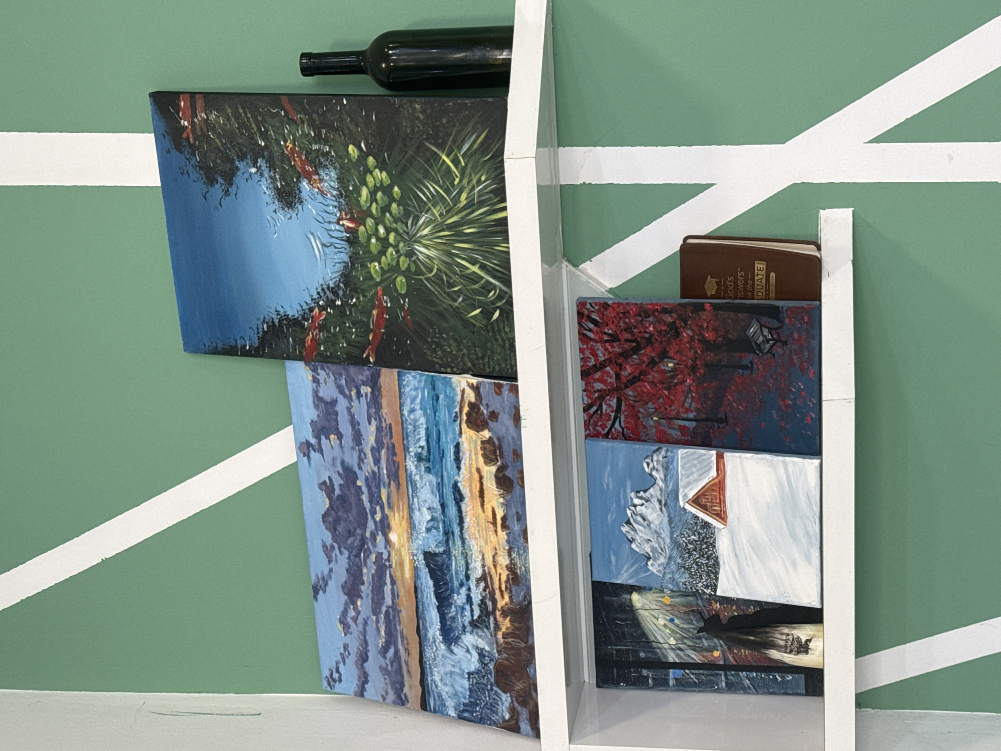 The image displays an arrangement of small landscape paintings on white shelves against a distinctive green wall. The green wall is marked with broad white diagonal lines, resembling the boundaries of a sports court.
On the upper left section, a dark green or black wine bottle rests horizontally above two paintings. The topmost painting depicts a tranquil pond with vibrant blue water, orange koi fish, and lush green aquatic plants, including lily pads and taller grasses. Below this, a painting showcases a dynamic seascape or beach scene during sunset or sunrise, with foamy waves breaking on the shore under a sky painted with blues, oranges, and purples.
On the right side, another set of white shelves holds three more paintings and a small book. The uppermost painting here features an autumnal setting with vivid red foliage dominating the scene, accompanied by a street lamp and possibly a bench. Below it, a snowy mountain landscape is depicted, featuring jagged white peaks and a red-roofed house or cabin. The bottom-most painting captures a dark night street scene, illuminated by bright streetlights casting reflections on a wet surface, possibly with faint figures.
A small, dark brown book is tucked onto the right shelf, with partial text visible on its cover: "God's Answers for the GRADUATE" and "on Wise". The collection of paintings presents various natural and urban scenes, ranging from serene ponds to dramatic seascapes and snowy mountains to city nights. The unique backdrop suggests an unconventional display setting, possibly within a themed space or a creative studio in Batumi, Georgia.