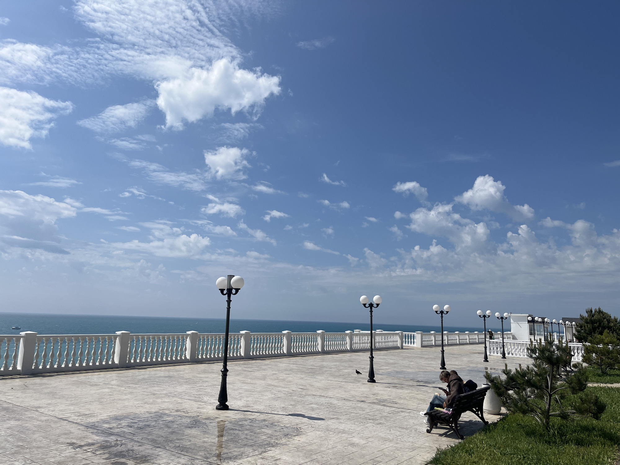 The image depicts a sunny day at a promenade in Anapa, Russia. The scene features a light grey concrete platform with a decorative white balustrade overlooking the sea. Several ornate lampposts are spaced along the promenade. A couple is sitting on a bench on the right side of the image, seemingly occupied with their mobile devices. A small bird stands on the ground nearby. The blue sea stretches to the horizon, with a ship visible in the distance. The sky is blue with scattered white clouds. On the right, there is a patch of green grass and small pine trees. A building with a white facade is visible behind the trees.