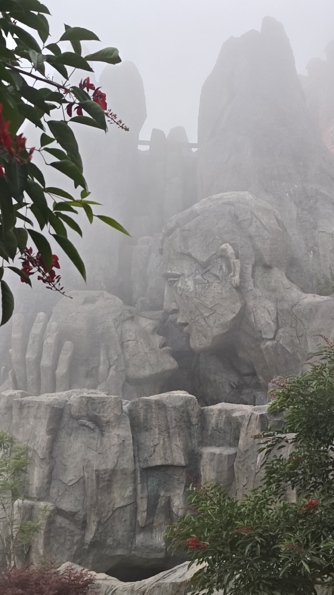 This image captures a misty, atmospheric scene in Đà Nẵng, Vietnam, showcasing a monumental rock sculpture. The primary subjects are two giant, carved stone faces, one male and one female, positioned intimately as if in a tender embrace or about to kiss. A large, sculpted hand appears to cradle the female face, emphasizing the gesture of affection.
The sculptures are intricately detailed with craggy textures, mimicking natural rock, and are integrated into a larger, rugged landscape of what appears to be massive rock formations or constructed cliff faces. The dominant color palette is various shades of gray from the stone, contrasting with the vibrant green foliage and clusters of small red flowers visible in the foreground and around the base of the sculptures.
The entire scene is enveloped in a thick fog or mist, which creates a soft, diffused light and significantly reduces visibility of distant elements. This misty weather lends an ethereal and romantic quality to the setting. In the background, indistinct outlines of more rock structures or mountains can be faintly discerned through the haze, suggesting a grand, elevated location.
The time of day is not clearly indicated due to the heavy mist, but the soft lighting suggests an overcast condition. There is no visible text in the image. The sculptures convey a powerful sense of love, intimacy, and grandeur within a dramatic, fog-shrouded environment.