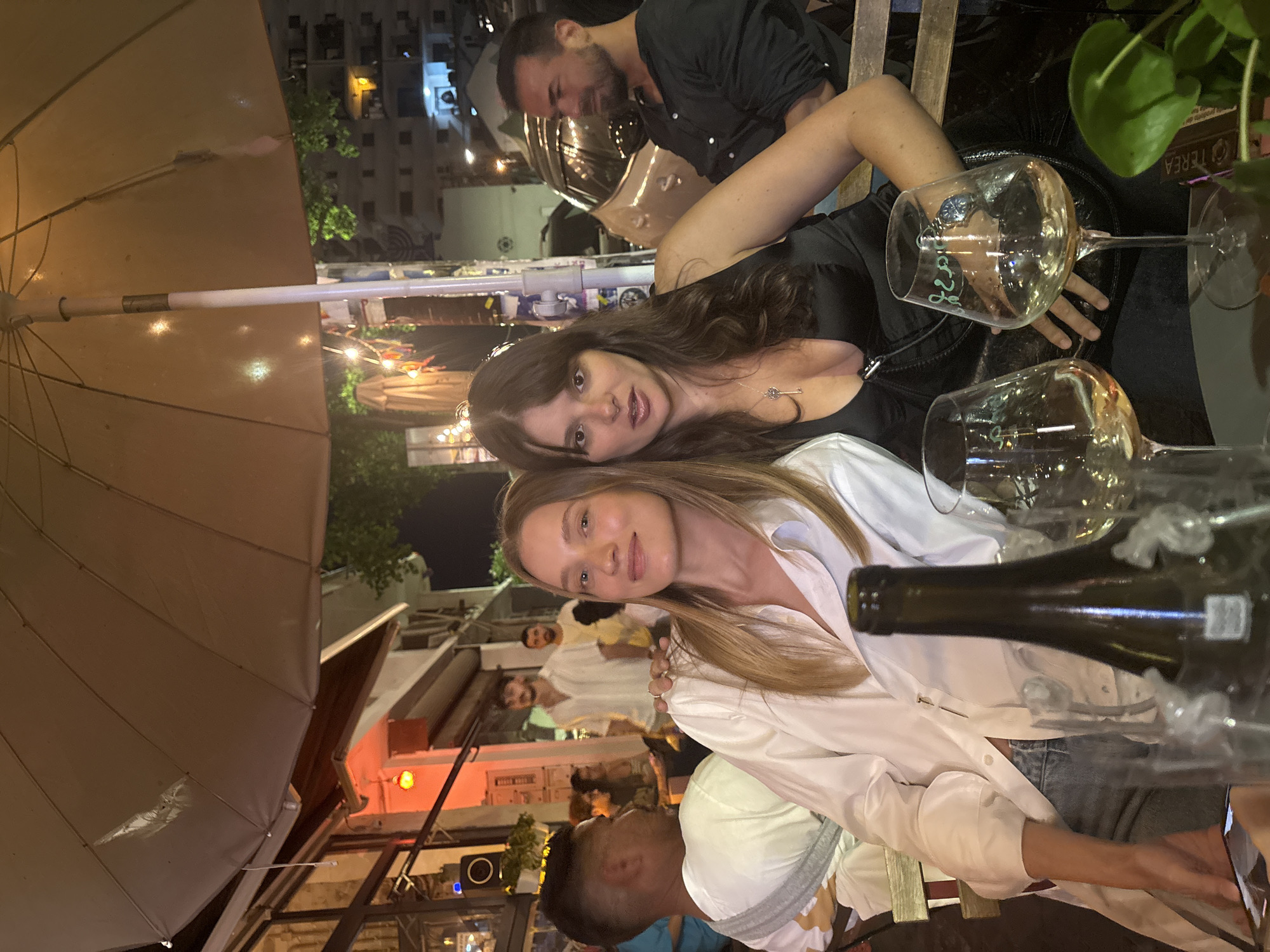 The photograph captures an outdoor evening scene in Tel-Aviv, Israel, likely a restaurant or bar patio, beneath a large umbrella providing shade. Two young women are seated at a table, posing for the camera. The woman on the left, with blonde hair, wears a white button-down shirt and blue jeans. The woman on the right, with dark hair, is dressed in a black satin top and a necklace with a key pendant. In the foreground, a bottle of wine and two wine glasses are visible on the table. In the background, other patrons and buildings are faintly discernible, illuminated by ambient lighting, including string lights and streetlights, suggesting a lively urban environment.