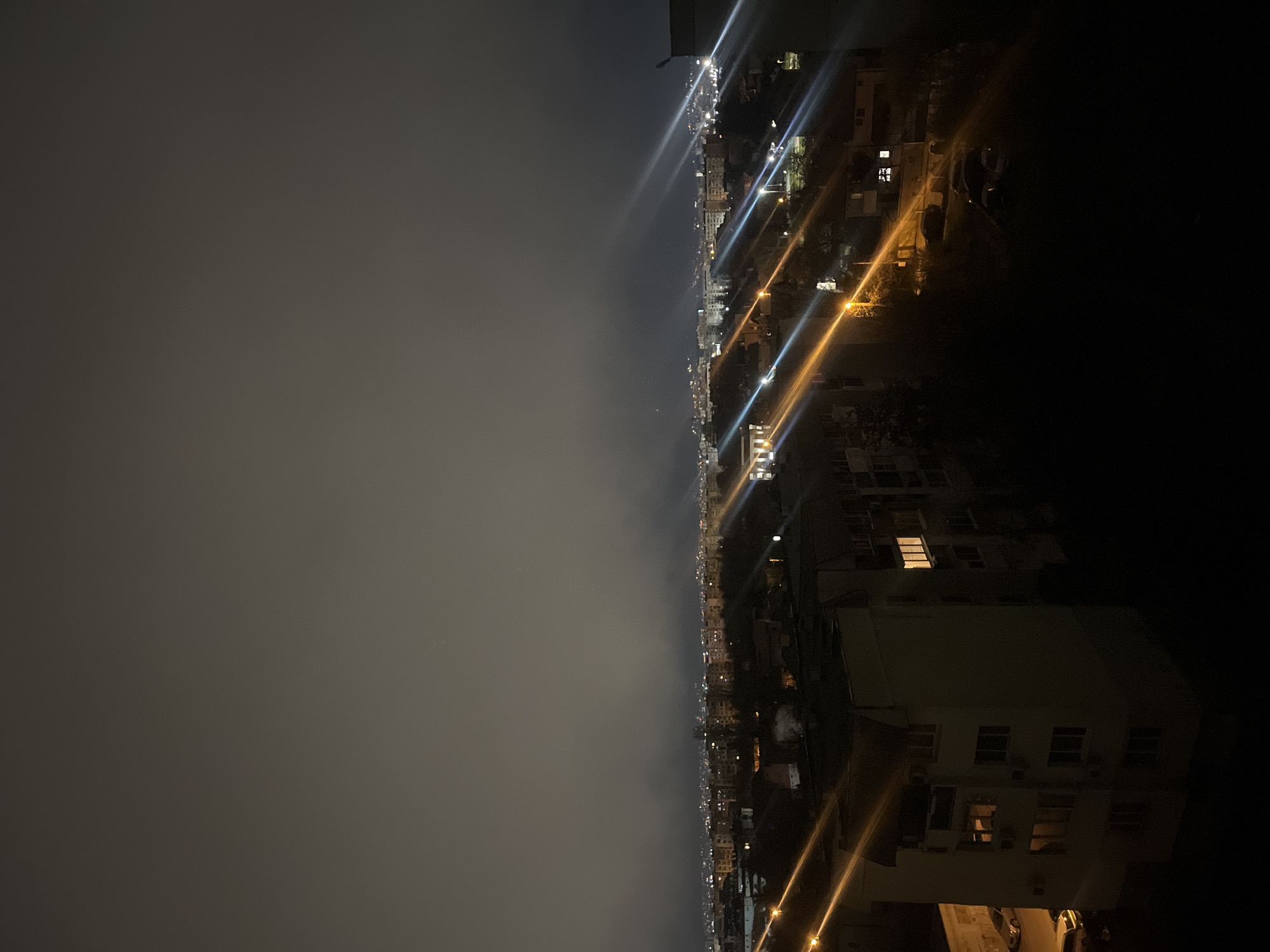 The image depicts an aerial view of a cityscape at night. A heavy fog or smog hangs over the left side of the frame, obscuring much of the city in that direction. The right side is more clearly defined, showing buildings, streets, and lights. The lights form lines, possibly indicating roads or avenues. There is no visible presence of people. Specific interactions or actions are unobservable due to the darkness and fog.
I can only make a reasonable assumption that the location is the city of Sofia, Bulgaria based on the prompt, not the image itself. I cannot determine the location based on the available image data.