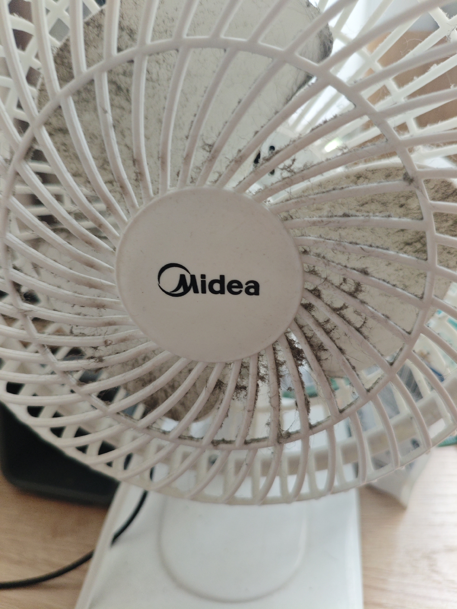 A close-up, slightly angled shot shows a dirty white Midea brand fan. The fan's grill, made of white plastic bars, is heavily covered in dust and debris, creating a gray and brown appearance. The Midea logo, a black stylized "M" within a circle, is visible on the center of the fan's protective grill. The fan is positioned on a white base, and a black power cord extends out of frame. The background suggests an indoor setting with a wooden surface visible. The image is taken from a perspective that emphasizes the fan's front. There are no people visible in the image. This image is located in Berdsk municipality, Russia.