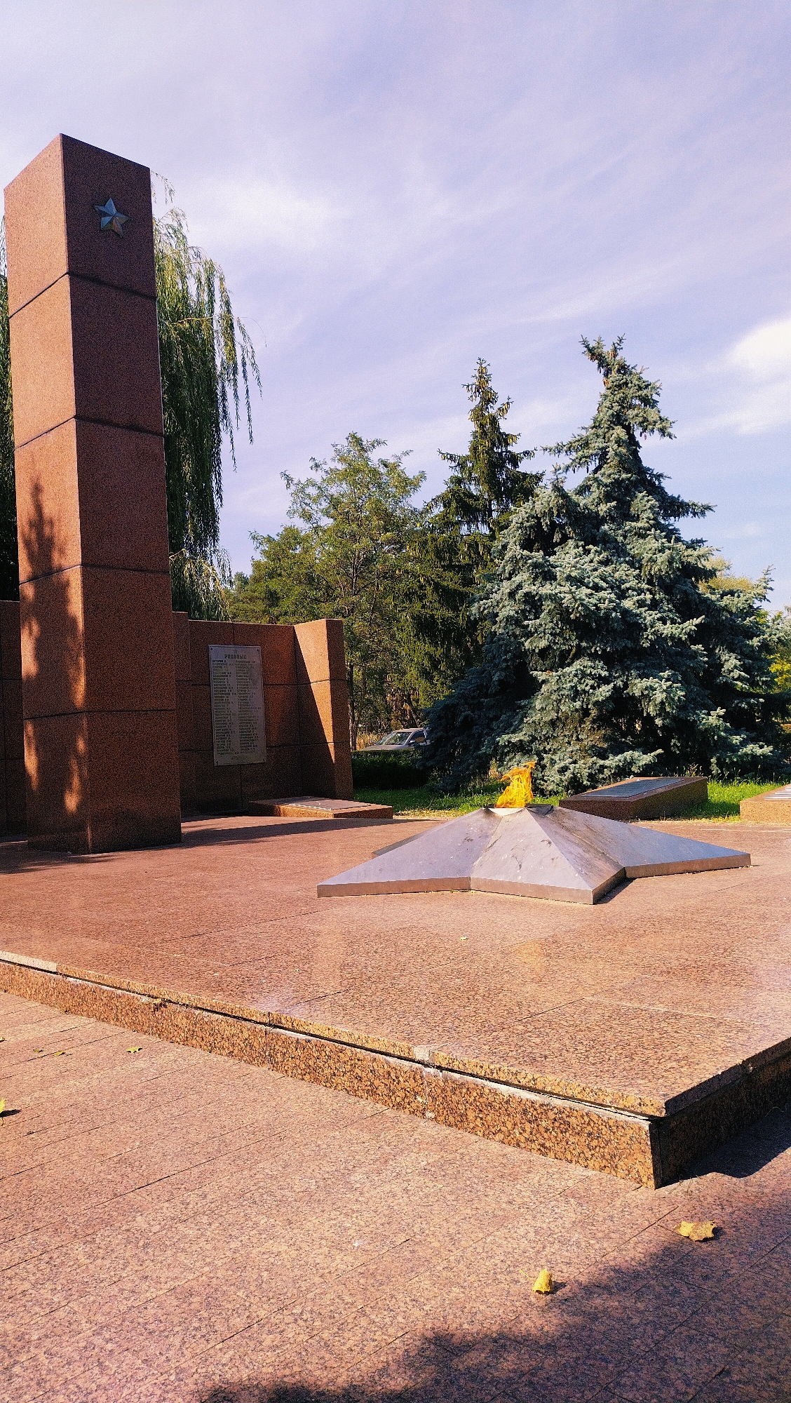 The image depicts a war memorial in a park-like setting, featuring a tall, reddish-brown stone obelisk with a star emblem at its apex. To the right of the obelisk, a star-shaped structure with a perpetually burning flame is prominent. Behind these structures, a variety of trees, including tall evergreen trees and a weeping willow, create a natural backdrop. The ground in front of the memorial is paved with reddish-brown tiles. A plaque is affixed to a section of the memorial near the obelisk. The sky above is partly cloudy with a blue hue. The lighting suggests it is daytime, with shadows cast by the obelisk and trees. There are no people visible in the scene.