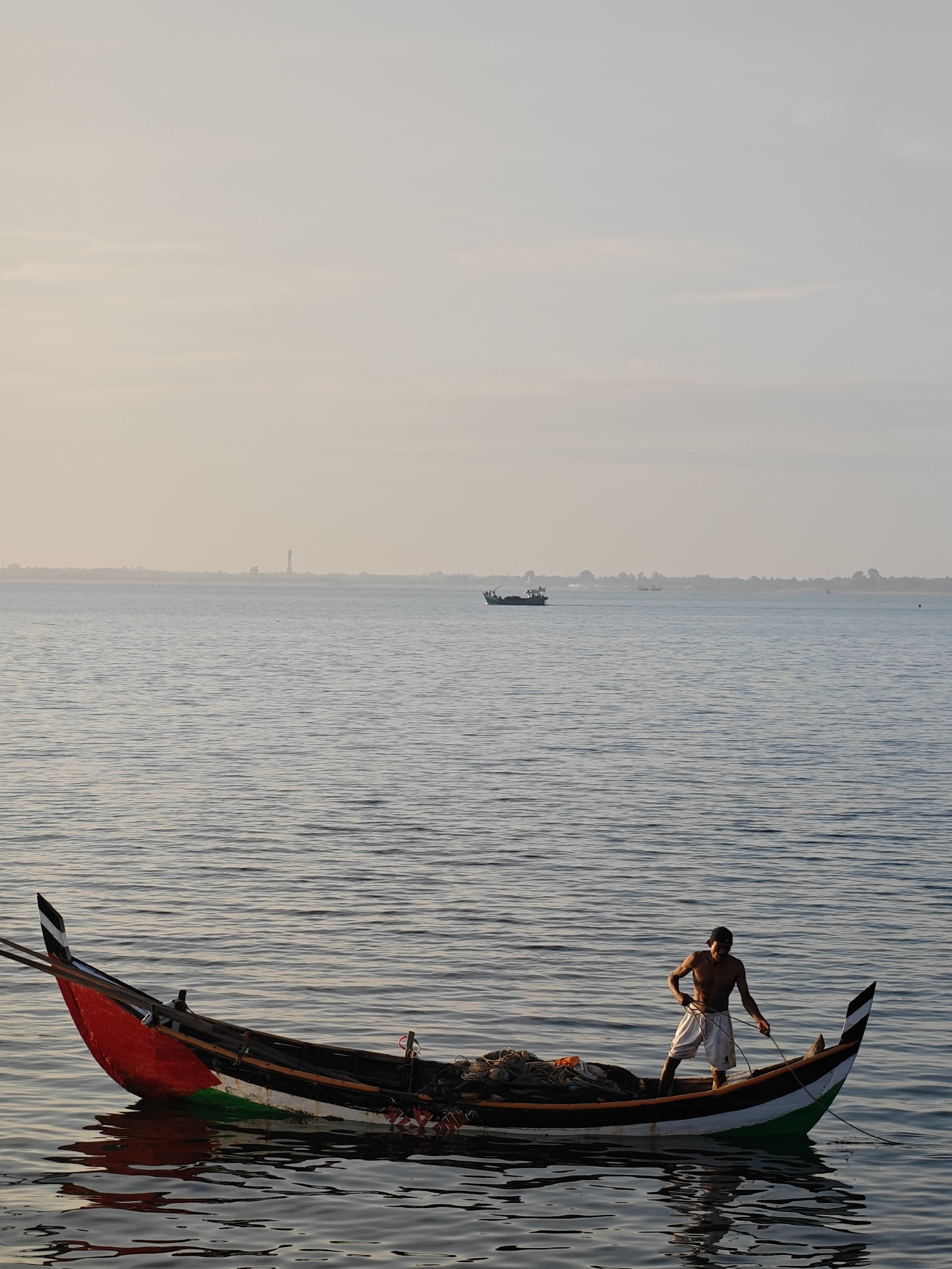 A man stands in a traditional wooden fishing boat, which is partially submerged in calm, rippled water. The boat is long and narrow, with a pointed bow and stern, painted red, white, and green. The man, who is shirtless and wearing white shorts and a dark cap, is holding onto a rope. The boat is laden with fishing nets. In the background, the sea stretches to the horizon, with a hazy sky above. A distant lighthouse and other boats are visible on the horizon. The scene suggests a fishing activity in the waters of Lhokseumawe, Indonesia, likely during dawn or dusk given the soft lighting.