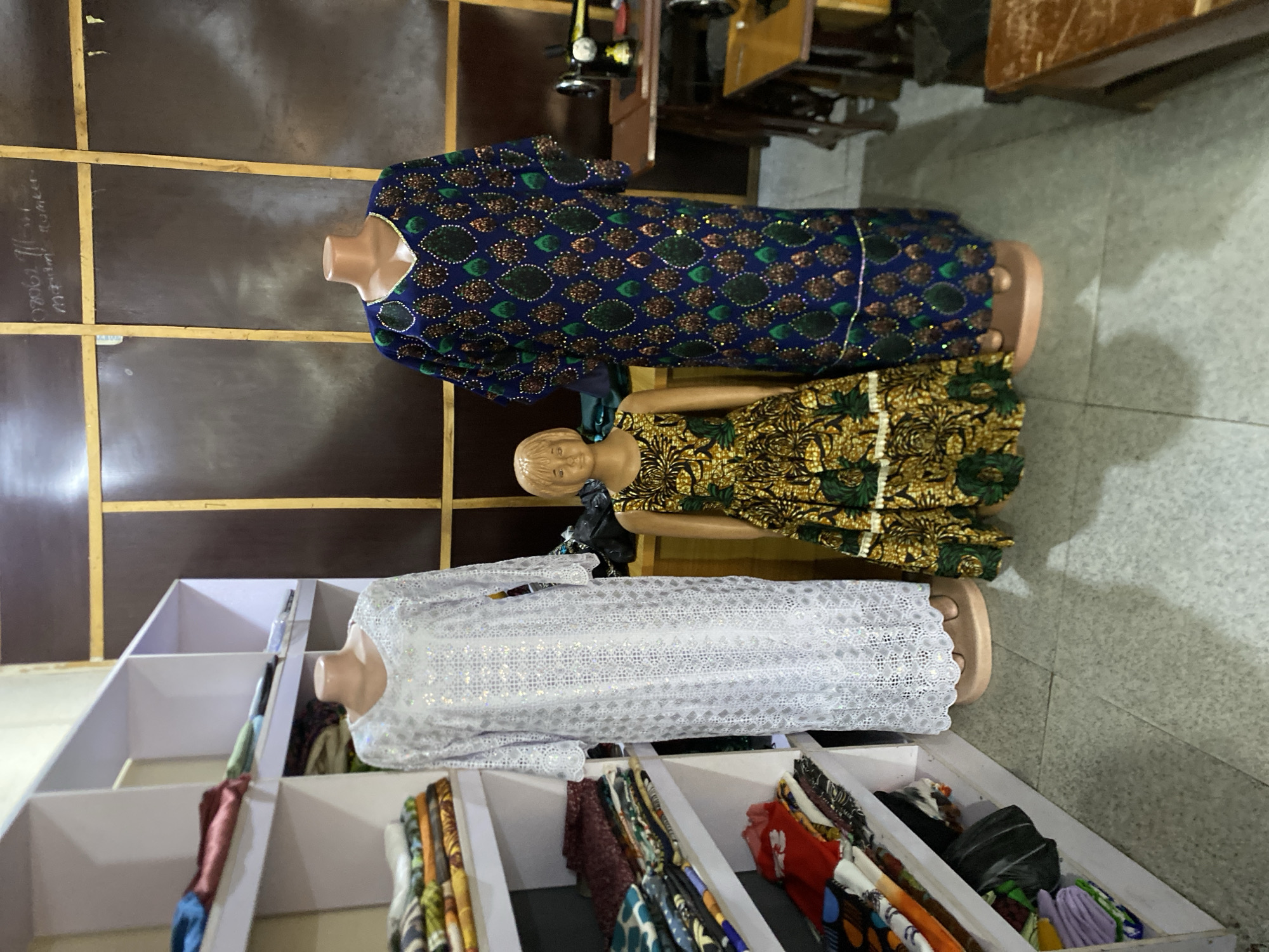 The image shows the interior of a clothing shop in Bukuru, Nigeria. Three mannequins are displaying different styles of women's clothing. The first mannequin, positioned at the top, is wearing a long-sleeved blue dress with green and gold detailing. Below it, a second mannequin is adorned with a short, patterned dress featuring shades of green and gold. The third mannequin at the bottom showcases a white, lace-like dress. Fabric rolls of various colors and patterns are visible in a display shelf near the white dress. A wooden wall with written notes is in the background. The floor appears to be tiled in a light gray color. Other furniture is also visible in the background.