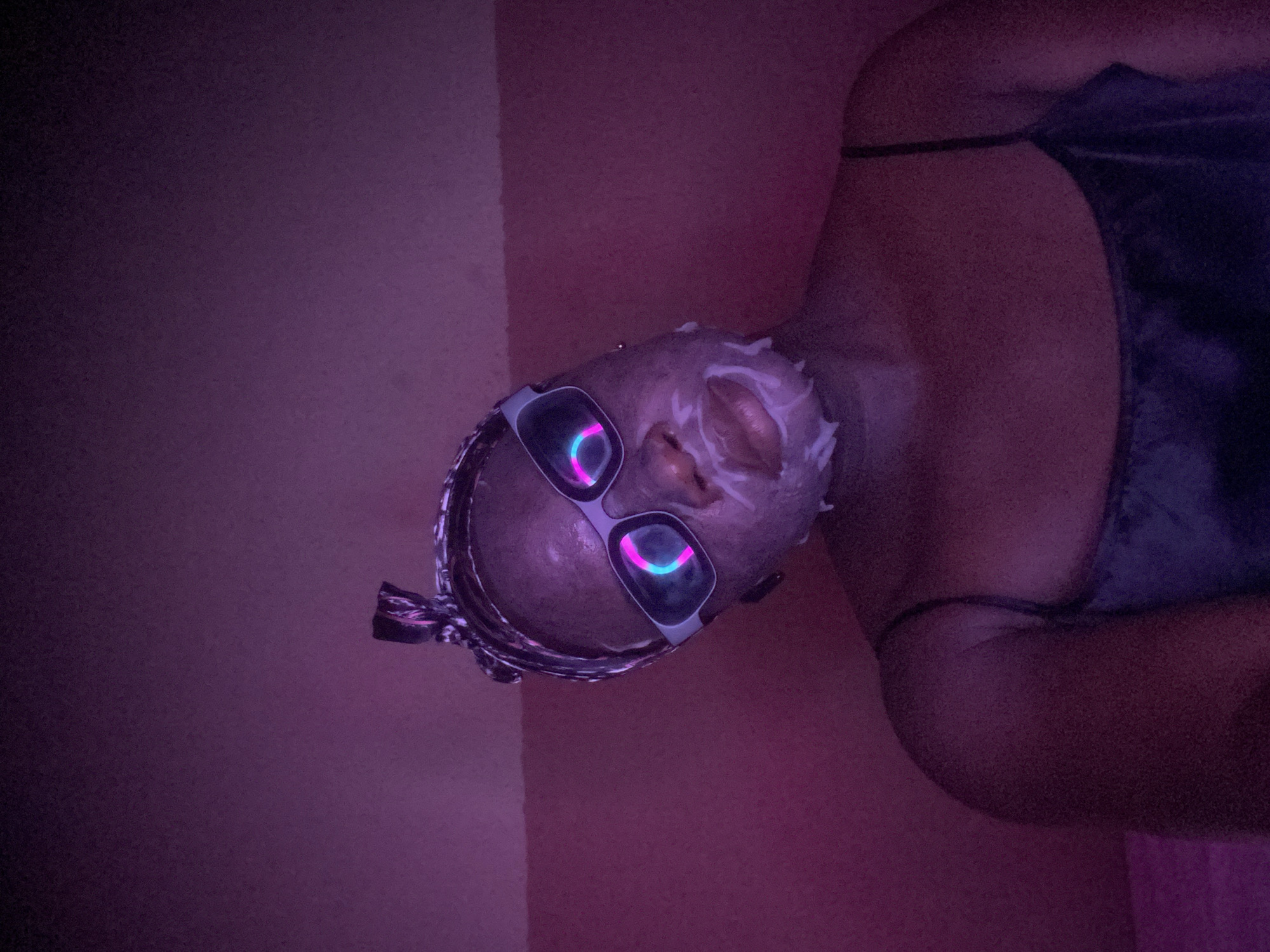 The image, taken in Maigizo, Nigeria, is oriented vertically, showing a person's head on the lower left and their torso extending upwards to the right. The scene is indoors and dimly lit with a predominant purple and pink ambient light, creating a relaxed or stylized atmosphere.
The primary subject is an individual with a white facial mask applied to their face, suggesting a self-care or beauty routine. They are wearing a pair of distinctive white-framed glasses with two glowing, multi-colored neon rings (pink and blue/cyan) visible through the lenses, adding a futuristic or playful element. The person's hair appears to be in braids or twists, secured back, possibly with a headwrap, though this is partially obscured by the glasses. They are wearing a dark-colored top, possibly made of velvet or satin, with a thin strap visible over the shoulder.
The background consists of a plain wall, reflecting the purple and pink lighting. The lighting suggests it is evening or night, or an intentionally darkened room with specialized lighting. No visible text is present in the image.