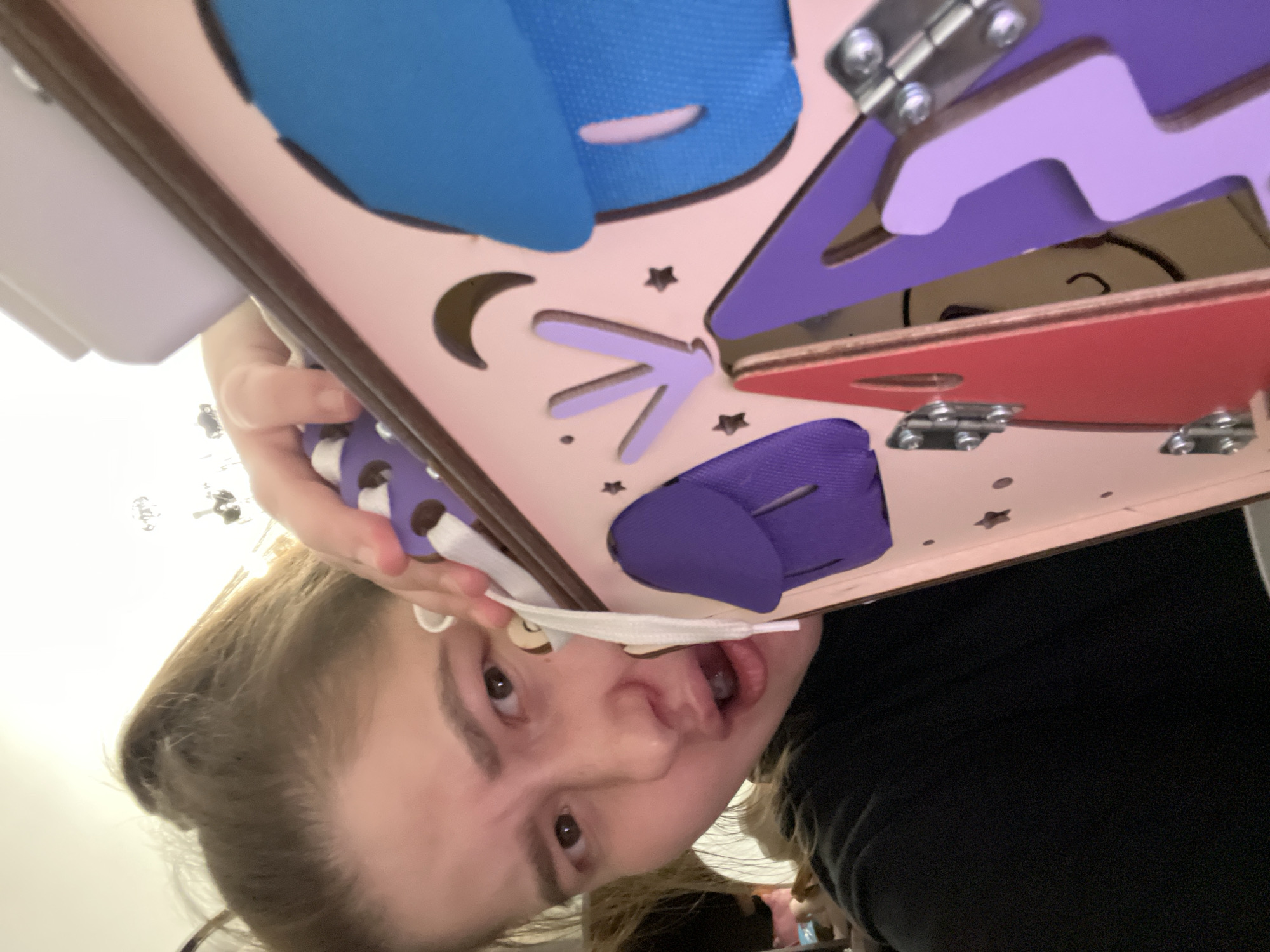 A young woman is holding up a busy board. The busy board is light pink with cut-out shapes including a moon, stars, and a geometric figure. On top of the busy board, there are several colorful fabric pieces, including blue, purple, and red. The fabric pieces appear to be fastened to the board with hinges, allowing them to be opened and closed. The woman is looking directly at the camera with her mouth slightly open. She has light brown hair pulled back, and she is wearing a dark shirt. The background is indistinct, showing a white wall and a few unidentified objects. The location is Unknown, Unknown.