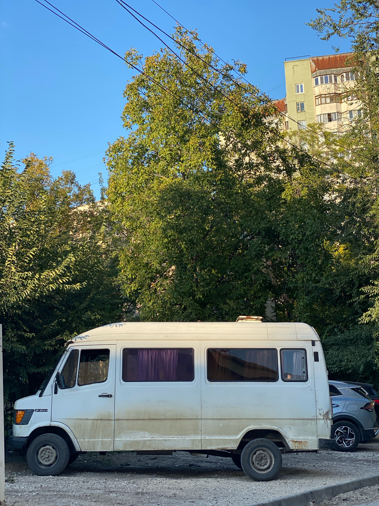 A white Mercedes-Benz 208D van is parked on a gravel surface beside a grey SUV. The van has visible dirt streaks along its lower body and some rust on its rear panel. Purple curtains are drawn behind the windows of the van. In the background, a light-green apartment building with multiple windows and a rusty roof can be seen behind lush green trees. Overhead, power lines stretch across a clear blue sky. This scene is likely located in Chișinău, Moldova, based on the architectural style and the presence of mature trees commonly found in urban areas of the city.
