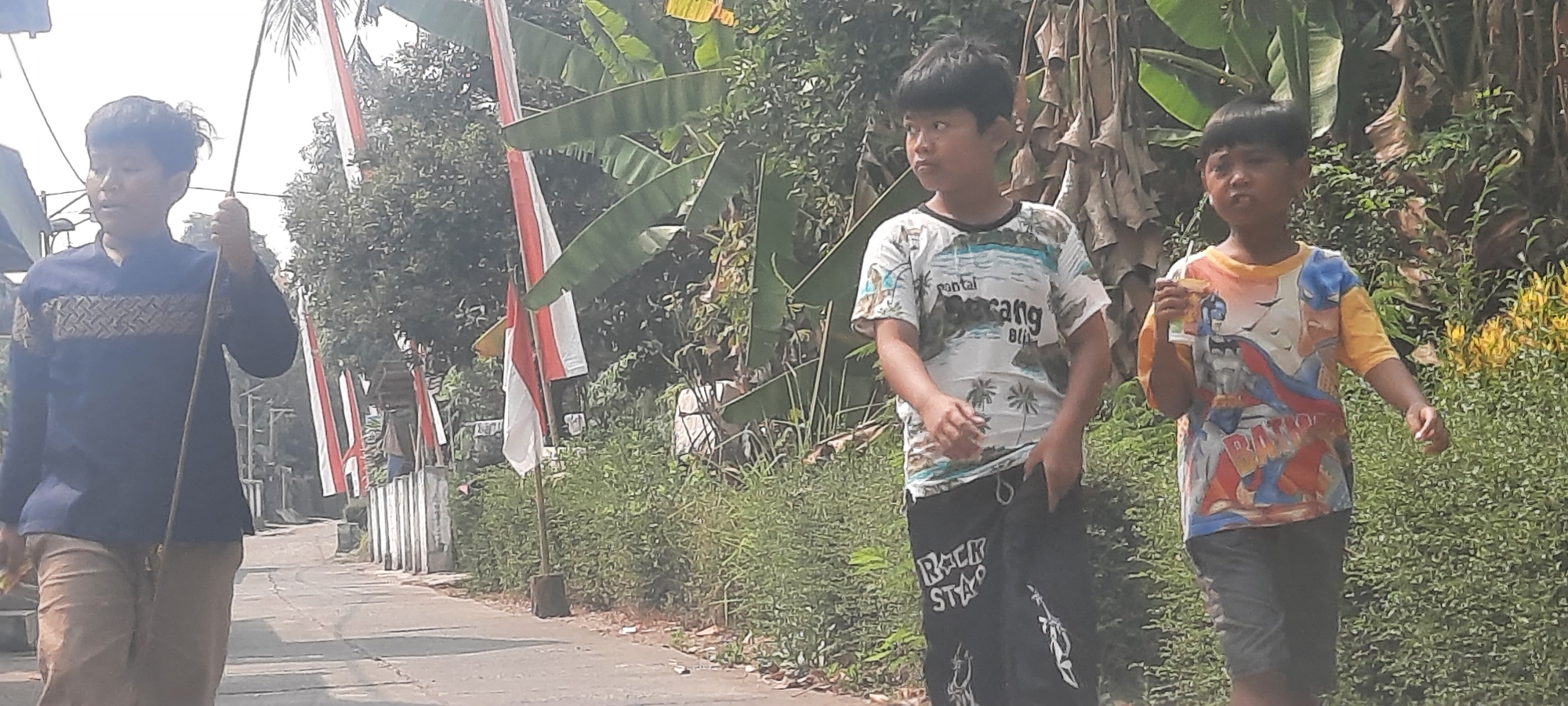 In Ploso, Indonesia, three young boys are walking down a paved road. The boy on the left wears a dark blue long-sleeved shirt with a geometric pattern on the chest and tan pants, and carries a long stick. The middle boy is dressed in a white t-shirt with a beach scene and palm trees, and black pants with "Rock Star" printed on them. The boy on the right wears a colorful t-shirt featuring Batman and yellow sleeves, and dark shorts, holding a beverage in a cup with a straw. Red and white Indonesian flags are visible on poles lining the road, and lush green vegetation, including banana leaves, surrounds the scene. The sky is bright and hazy.