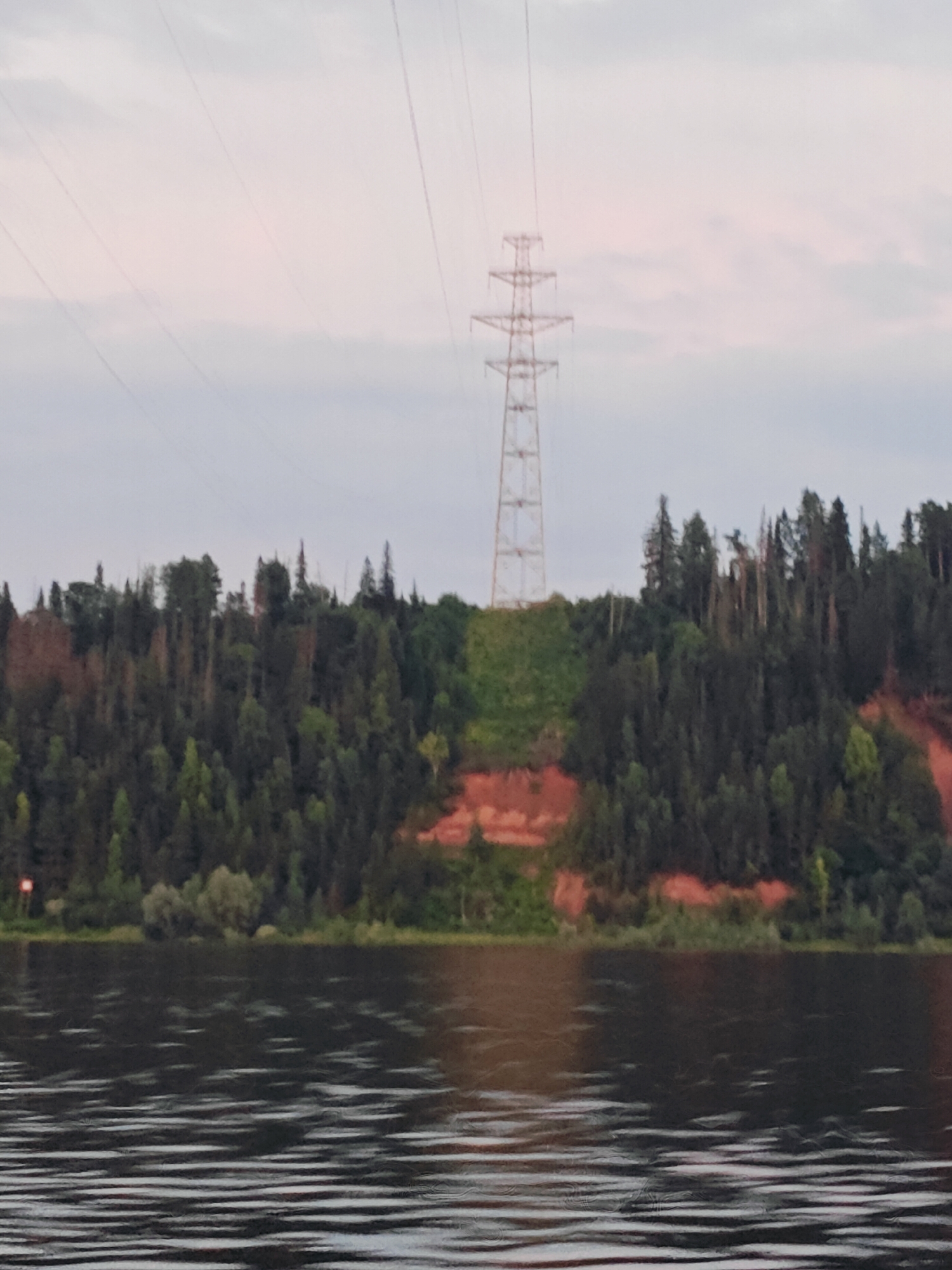 This is a medium-shot, eye-level, outdoor photograph of a wooded hill with a power line tower visible against a hazy sky. The hill is covered in a dense forest of various shades of green trees. Reddish-brown earth is exposed on a section of the hillside, creating a diagonal path. A large, metallic power line tower stands prominently on the left side of the hill, with wires extending across the frame. Below the hill, a dark body of water with ripples is visible. The sky is a light gray with some faint pinkish hues. The scene appears to be a natural landscape with industrial infrastructure. The photograph was taken in the city of Noviy, Russia. There are no people or animals present in the image.
