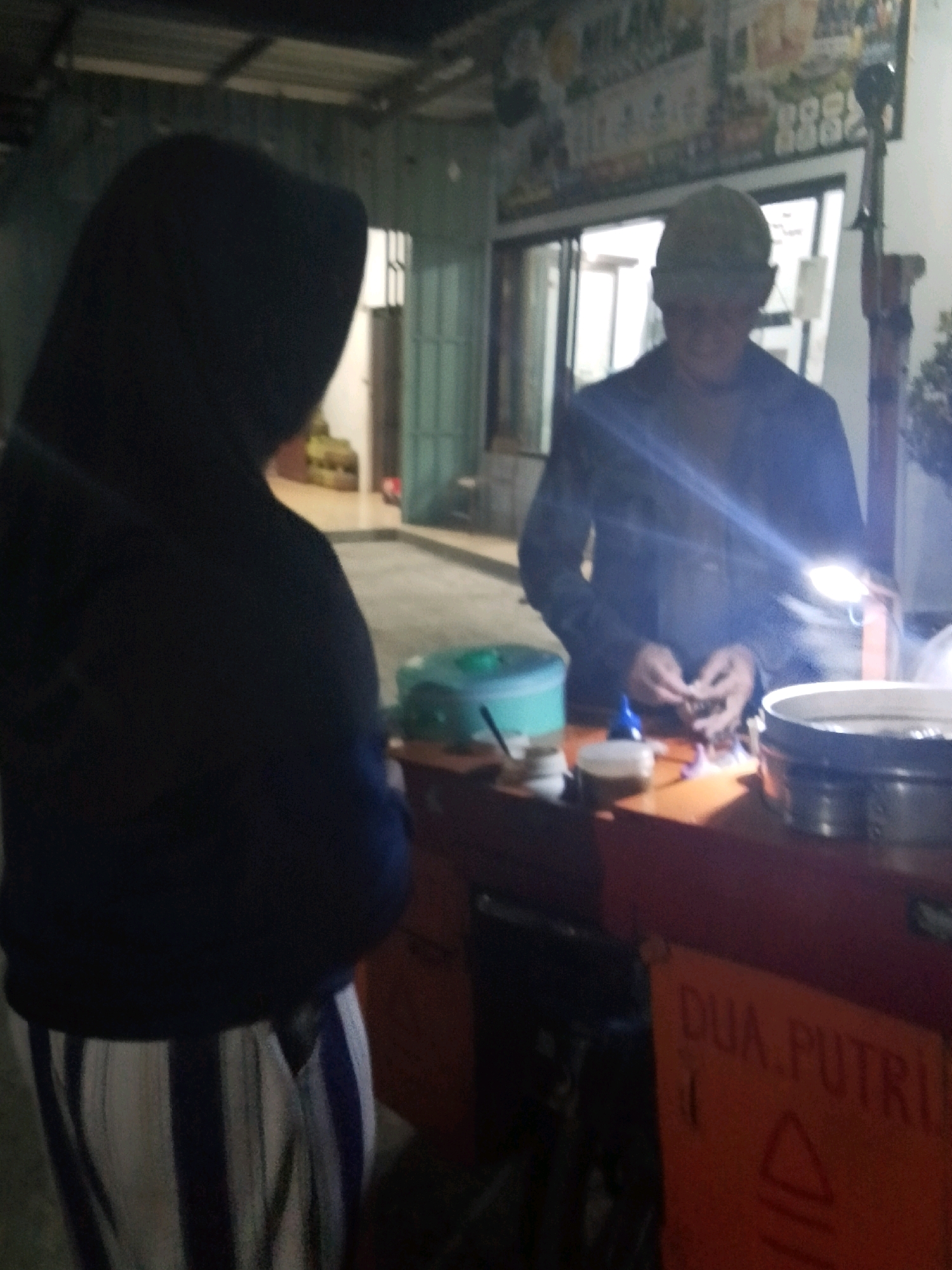 The image depicts a dimly lit outdoor scene, likely a food stall, in Bulurejo, Indonesia. In the foreground, a person wearing a dark hooded jacket and vertically striped pants (white and blue) stands facing a stall. Behind them, to the right, a second person wearing a dark jacket and a light-colored cap is actively engaged in preparing something at the stall. This person is illuminated by a bright light source, possibly a flashlight held in their hand. The stall itself is orange and features a sign with the text "DUA. PUTRI" and a triangle symbol. On the stall, several containers and items are visible, including a light blue bin, small jars, and a large metal basin. The background suggests a somewhat rustic or informal setting with a corrugated metal roof visible overhead and buildings or structures in the distance. The overall atmosphere is that of a casual, evening transaction at a street-side vendor.