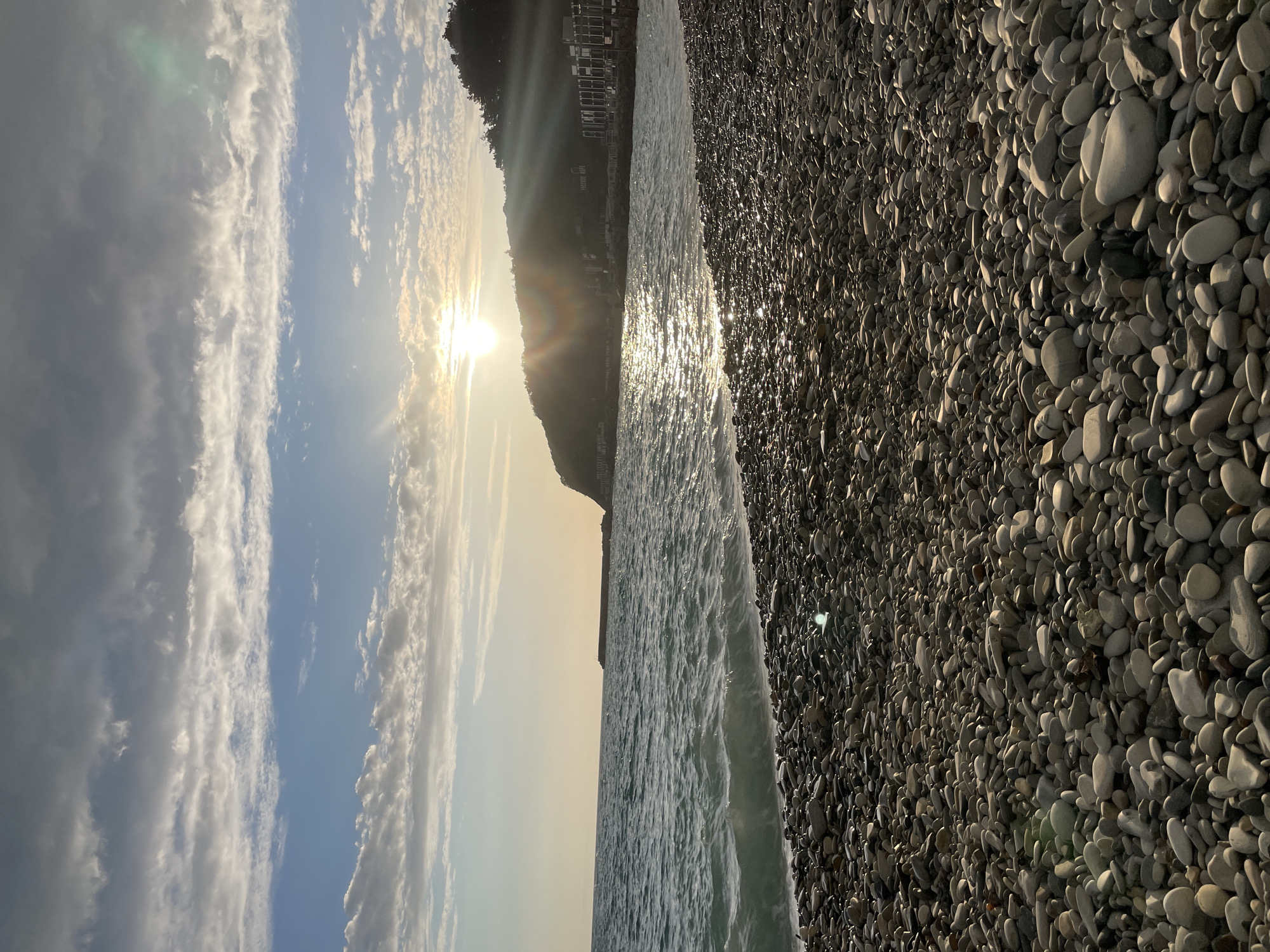 The image depicts a beach scene in Olginka, Russia, at what appears to be sunset. A rocky shoreline filled with gray and white pebbles extends from the lower right towards the center. The sea, reflecting sunlight, occupies the central portion of the frame. The sun is partially obscured by a coastal mountain in the middle ground, with buildings visible on the hillside. The sky shows a gradient from blue on the left to a golden hue near the horizon, scattered with clouds.
