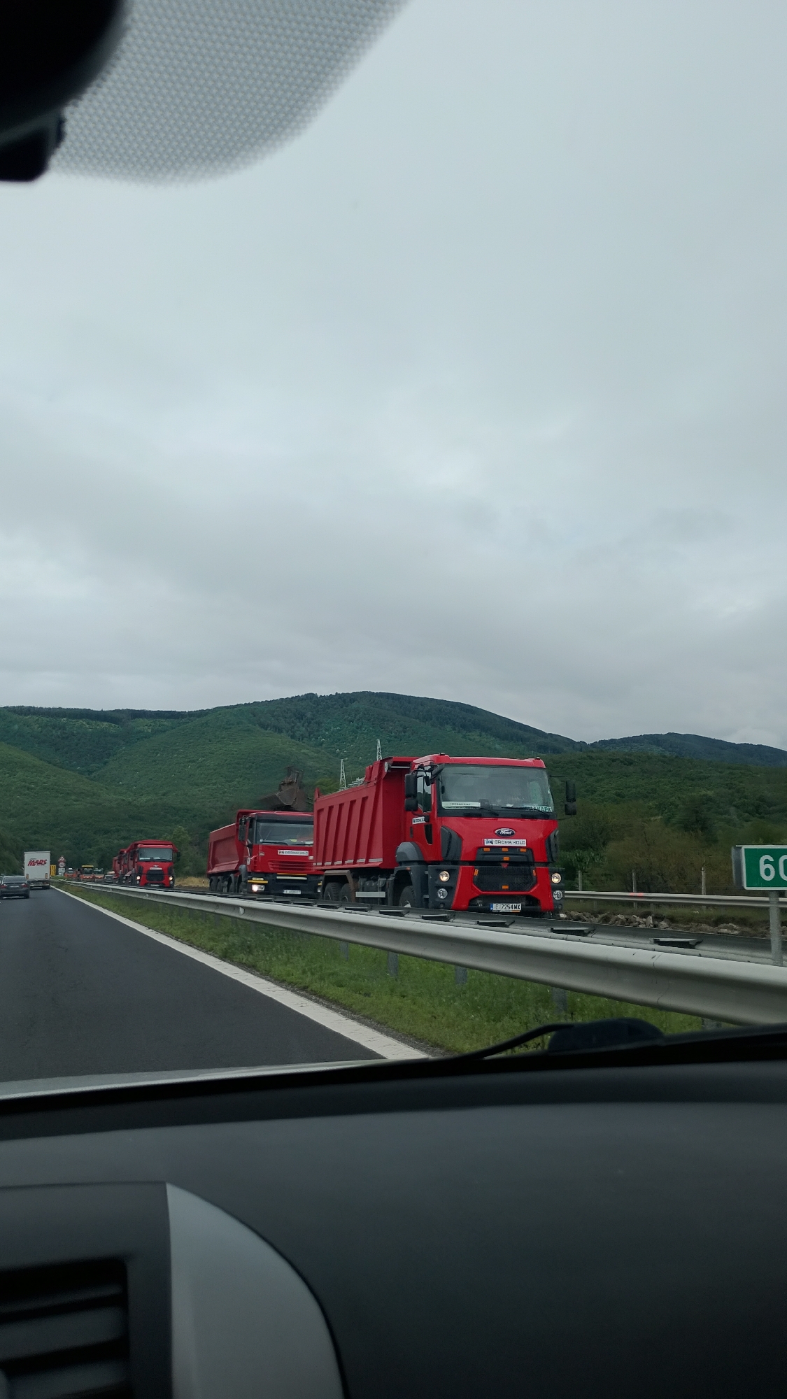 The image depicts a convoy of red dump trucks traveling on a highway under an overcast sky. The trucks, appearing to be Ford brand based on the emblem on the closest vehicle's grille, are in a line, suggesting a controlled movement. One of the trucks has a license plate that reads "E 7254 ML". The highway is bordered by a metal guardrail and a grassy shoulder. In the background, rolling green hills covered in trees suggest a rural or semi-rural setting. A speed limit sign indicating "60" is visible on the right side of the highway. The foreground shows a portion of the interior of a vehicle, likely a car, indicating the perspective of someone observing the convoy. The scene is consistent with a convoy moving through the region of Dolno Varshilo, Bulgaria, given the context of the prompt, though this specific location is not visually confirmed within the image itself.