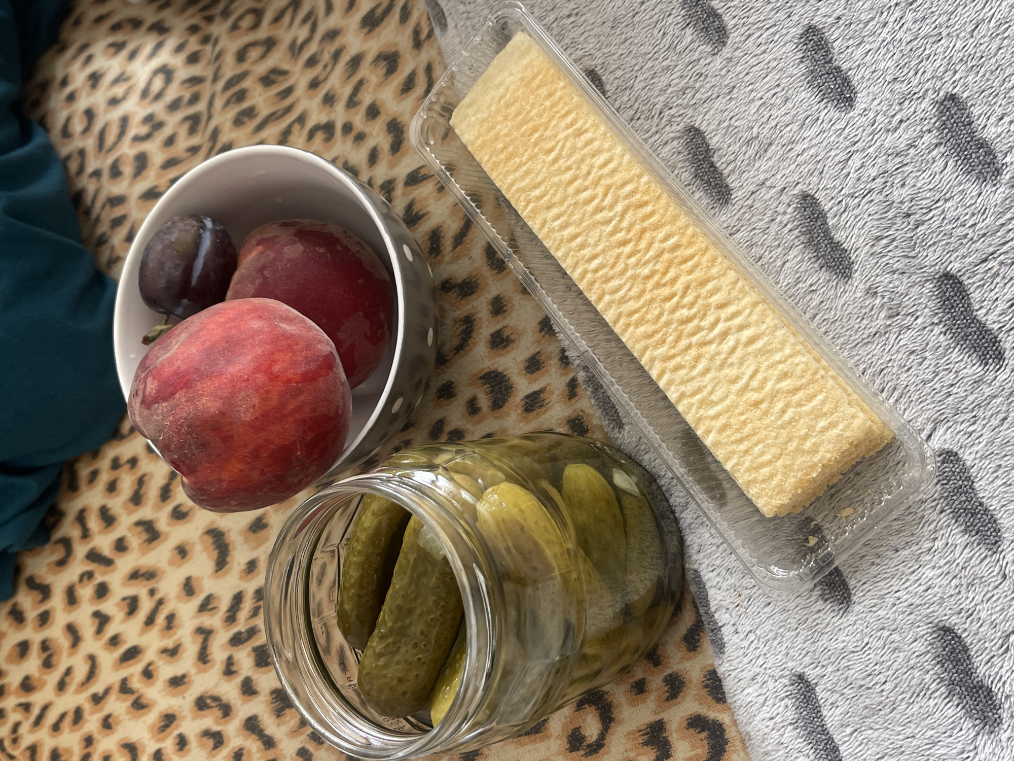 The image depicts a close-up, high-angle shot of various food items placed on a leopard-print fabric and a grey, textured blanket. In the foreground, a clear glass jar filled with pickles stands on the left. To the right of the jar, a rectangular, yellow biscuit is presented in a clear plastic container. In the background, a white bowl contains three pieces of fruit: two red peaches and one dark purple plum. A dark teal fabric is visible in the upper right corner. The overall setting is indoor, with no specific identifiable landmarks that would indicate a location in Varna, Bulgaria.