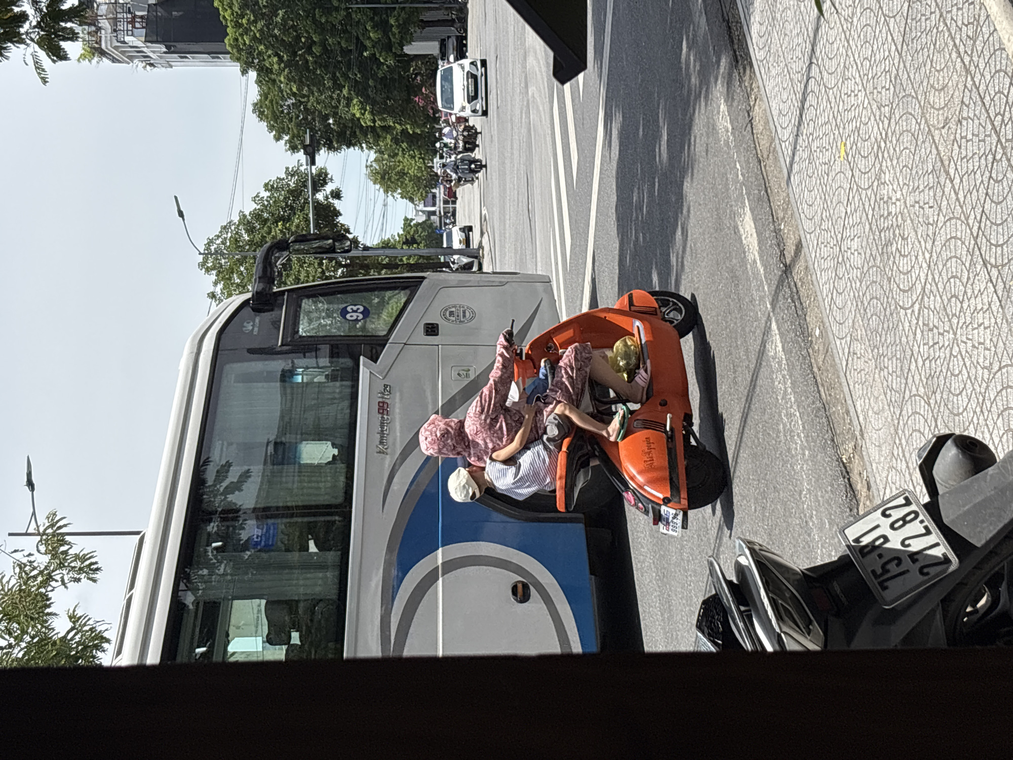 An orange electric scooter with two occupants, a woman and a child, is traveling on a street in Thuận Hóa Ward, Vietnam. The woman, wearing a pink patterned outfit and a headscarf, is operating the scooter. The child, wearing a striped shirt and a white cap, is seated in front of her. The scooter has a license plate that reads "99B 994.94".
A large white and blue bus is to the left of the scooter. Other vehicles, including motorcycles and cars, are visible on the street in the background, suggesting moderate traffic. The environment appears to be an urban setting with trees and buildings lining the street. The sky is bright, indicating daylight. The foreground shows a portion of another parked black motorcycle with a license plate that reads "75-B1 272.82". The street is paved and a pedestrian walkway with a patterned tile design is visible to the right.