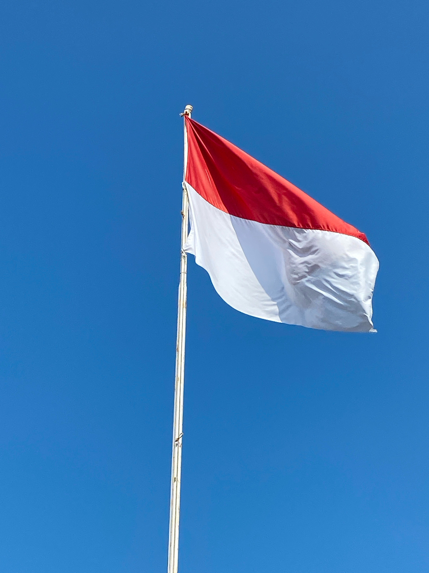 A national flag of Indonesia, red and white, flies from a tall white flagpole against a clear, bright blue sky. The flag is partially unfurled and billowing slightly in the wind, revealing its two horizontal stripes. The flagpole is positioned on the right side of the frame, with the flag extending towards the left and top. The background is a uniform expanse of blue sky, suggesting an outdoor setting with good visibility. The image appears to be a photograph taken from below, looking up at the flag. The location in the city of Karang Gading, Indonesia, is noted as part of the scene's context.