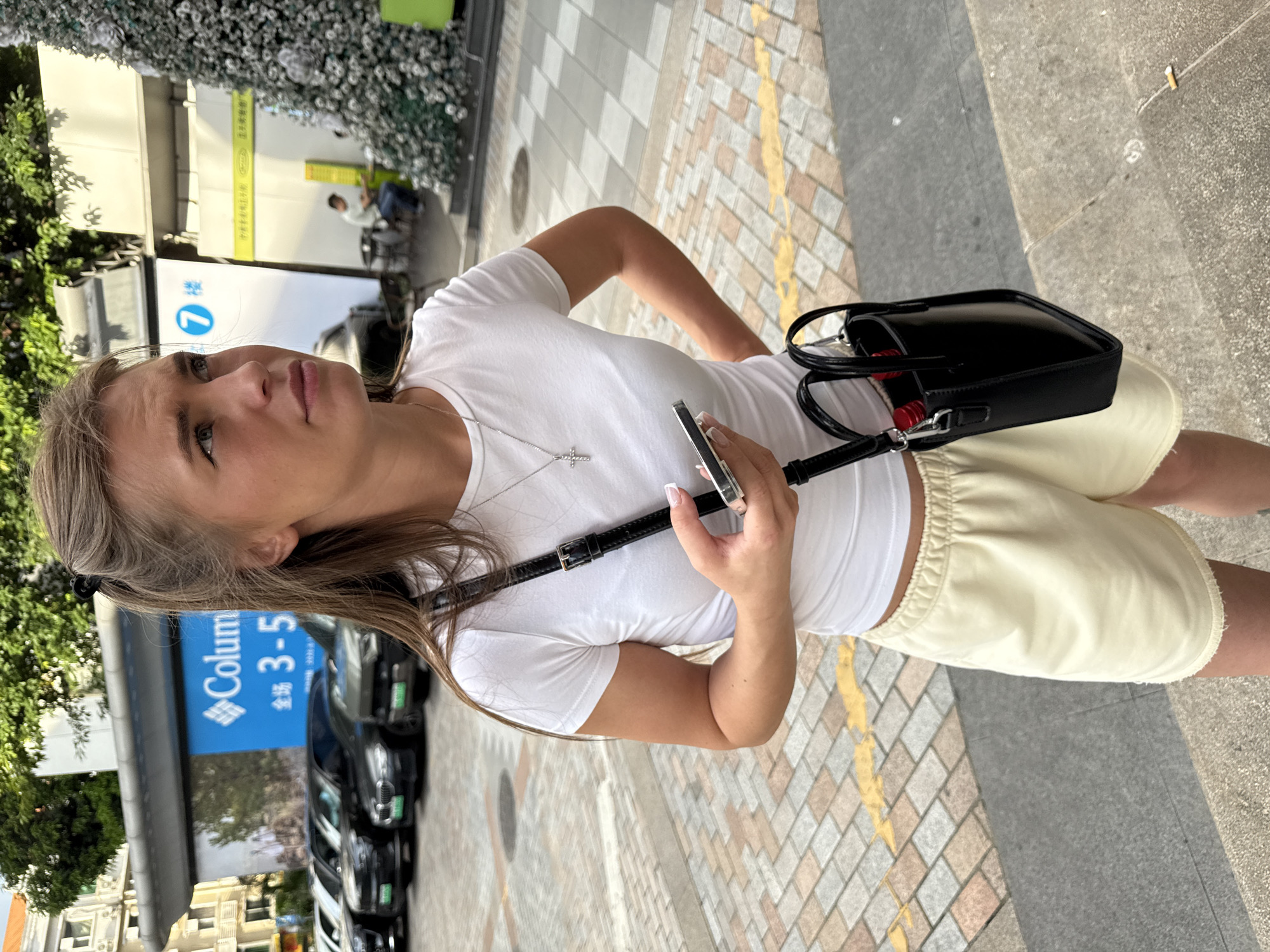 The image shows a young Caucasian woman standing outdoors in what appears to be a commercial area. She is wearing a white t-shirt, light-colored shorts, and a black crossbody bag. In her right hand, she holds a smartphone. Her hair is light brown and styled with a clip. Behind her, several black cars are parked in a row. A large billboard with blue and white text is visible in the background, along with a decorated Christmas tree on the right side of the frame. The ground is paved with rectangular tiles. The location is identified as Haizhu District, China.