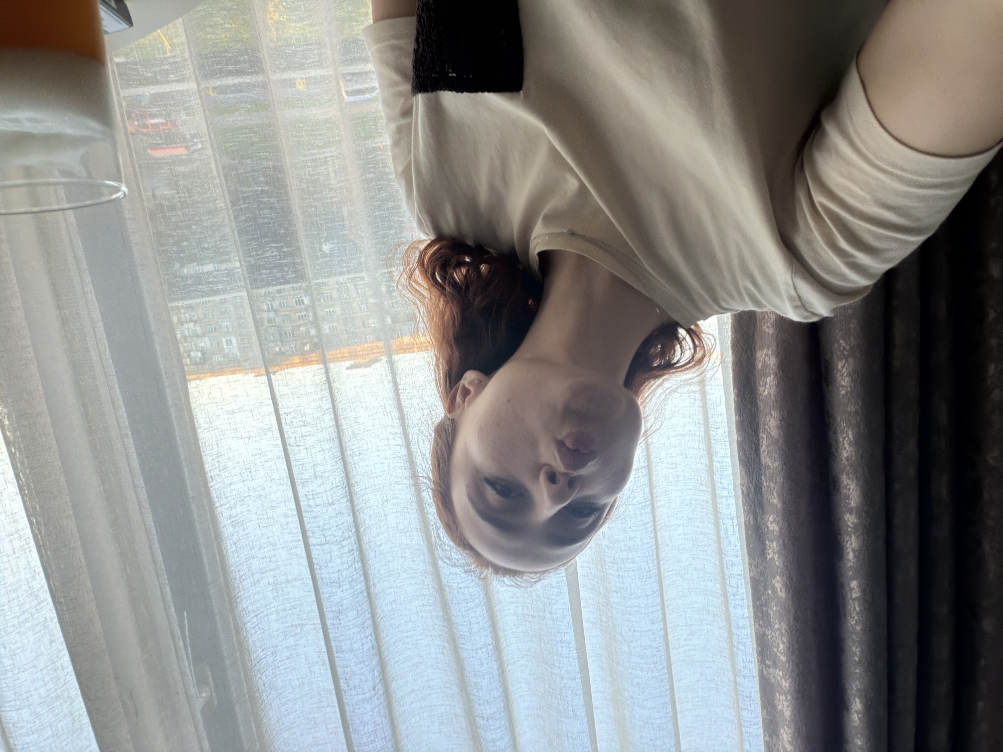 A young woman with red hair wears a white t-shirt with a dark patch on the shoulder. She is in a room with white curtains, and what appears to be gray patterned drapes on the right side of the frame. Outside the window is a view of Tbilisi, Georgia with apartment buildings. A glass, possibly containing liquid, is partially visible in the upper left corner.
