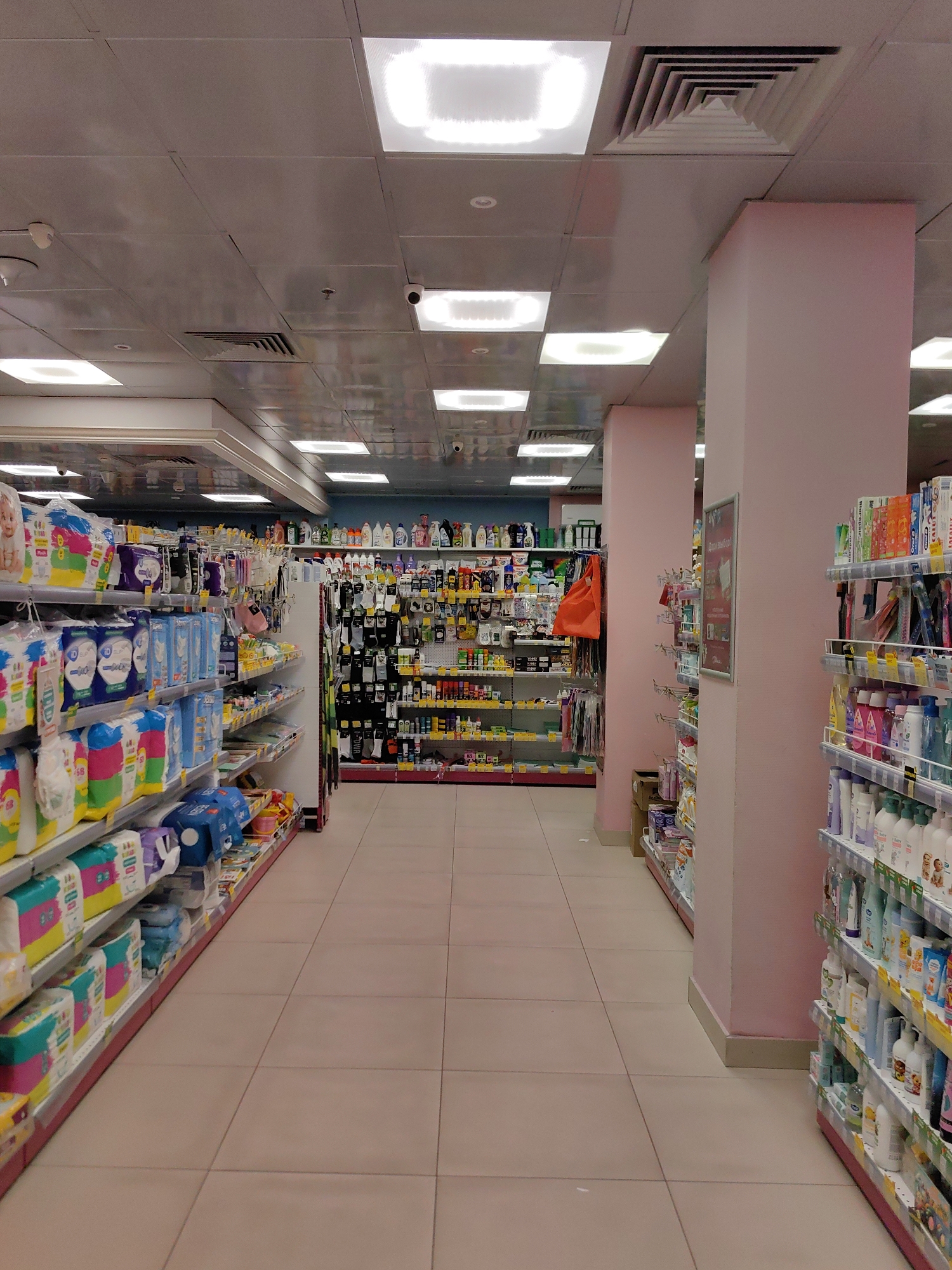 This is a medium-wide shot of an aisle in what appears to be a retail store, possibly a pharmacy or a supermarket, with products displayed on shelves. The aisle is clean and well-lit with recessed ceiling lights. On the left, there are shelves stocked with various packages of baby diapers and other baby care products. The products are neatly arranged in their original packaging. On the right, there are shelves displaying a variety of personal care items, including bottles of shampoo, lotion, and other toiletries. The shelves on the right are also well-stocked and organized. The floor is tiled in a light beige color. There are no people visible in the image. The environment is sterile and orderly, typical of a retail setting. The image appears to have been taken from a standing position, looking down the aisle. The store interior is modern and functional. Given the context of this being a specific location, and assuming this is a general store in Minsk, Belarus, the scene depicts a typical retail environment within the city.