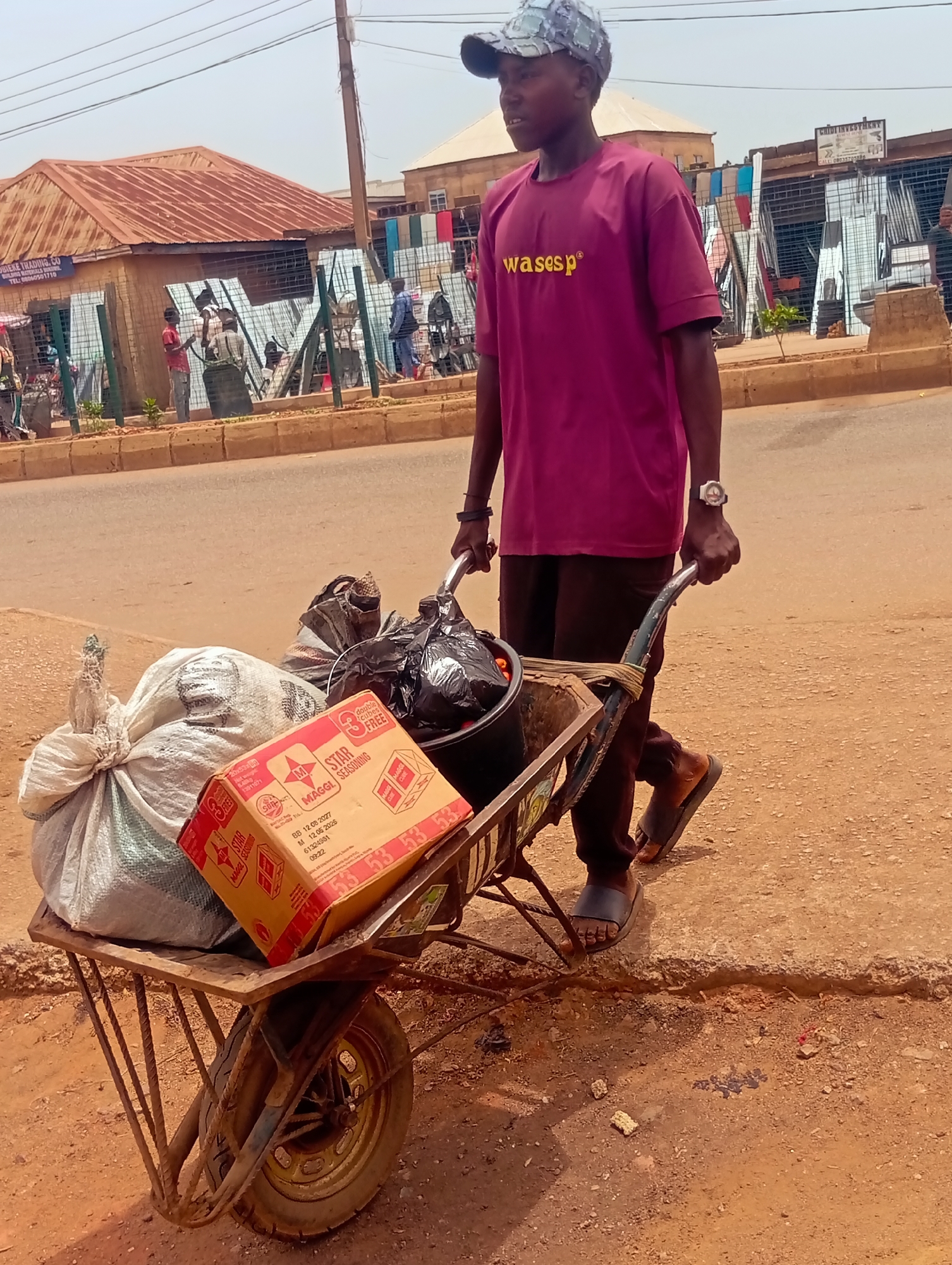 A man walks along a sidewalk in Bukuru, Nigeria, pulling a loaded wheelbarrow. He wears a magenta t-shirt with "wasesp" printed on it, brown trousers, and sandals, and has a patterned baseball cap on. He wears a watch on his left wrist. The wheelbarrow contains a large white sack, a box labeled "STAR MAGGI SEASONING," and black trash bags. A rusty roofed building is behind him with a sign that reads "OBIEKE TRADING CO". Other people are visible in the background, and a faded sign reads "CHIDI INVESTMENT" is also in the background. The environment appears to be a streetscape with mixed commercial and residential buildings.