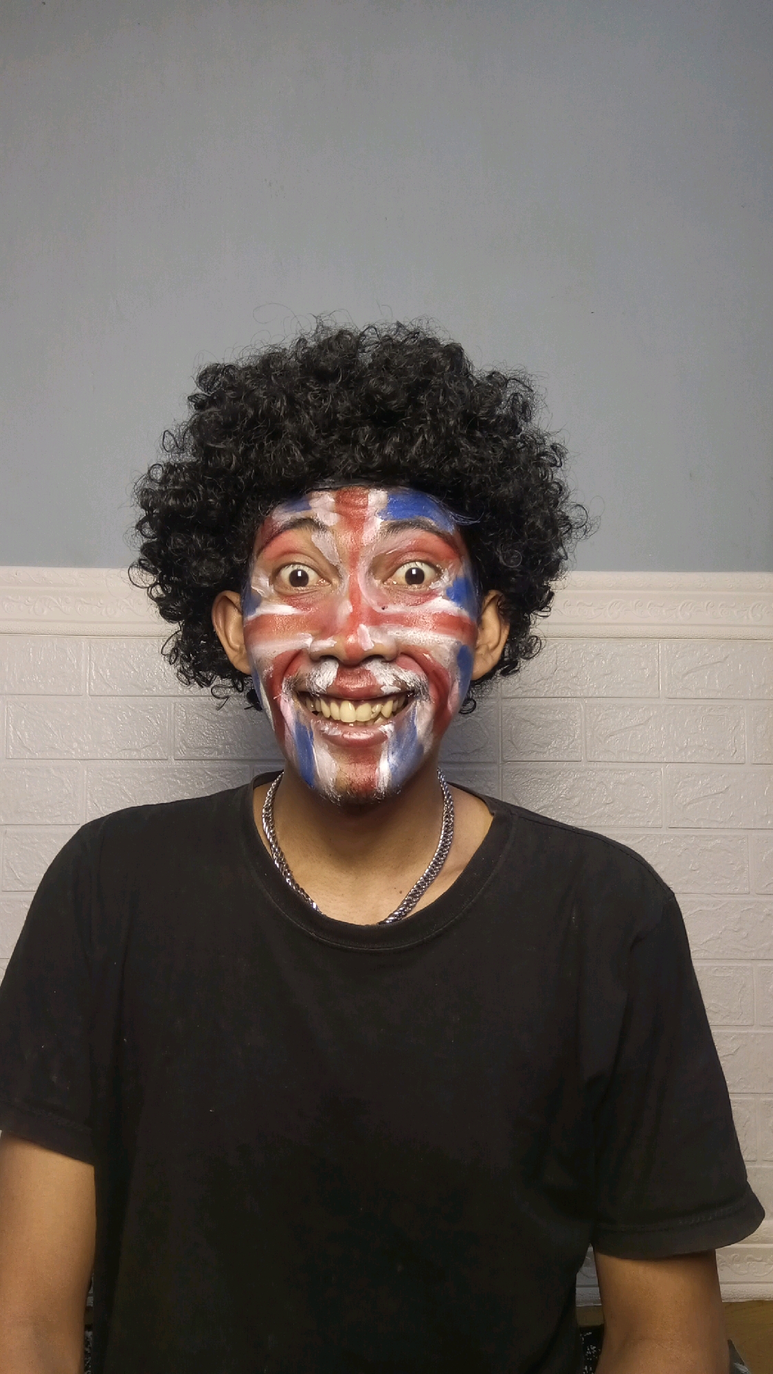 A person with a Union Jack flag painted on their face and a black afro wig is shown. They are wearing a black t-shirt and a silver chain. The background consists of a light grey wall with white textured panels. The image was taken in Kebumen, Indonesia.