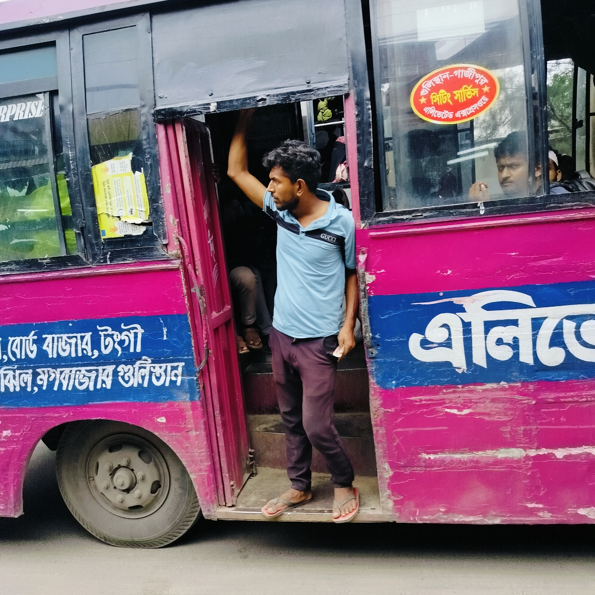 This media file captures a street scene, likely in Bangladesh, focusing on a bright pink and blue bus with prominent Bengali script. The bus appears to be in motion or stopped for passengers, as a man in a light blue polo shirt with "GUCCI" subtly displayed on it is standing in the open doorway, looking out. He is holding onto the doorframe with his left hand and appears to be either boarding or disembarking. His dark trousers and flip-flops suggest a casual, everyday setting.
Inside the bus, other passengers are visible, including a man in the front window who seems to be looking towards the viewer. Yellow posters with Bengali text are attached to the interior of the bus, and a red and yellow oval sticker with Bengali text is affixed to the window. The bus itself shows signs of wear and tear, with peeling paint contributing to a sense of realism and urban life.
The Bengali text on the side of the bus appears to indicate routes or destinations, with phrases like "বোর্ড বাজার, টংগী" (Board Bazar, Tongi) and "ঝিল, মগবাজার গুলিস্তান" (Jheel, Mogbazar Gulistan) visible. The text on the oval sticker translates to "Gulistan-Gajipur Seating Service, Elevated Expressway."
The overall impression is that of a typical public transportation scene in a bustling urban environment, capturing a moment of daily transit. The lighting suggests it could be daytime, with natural light illuminating the scene. The condition of the bus and the presence of the passengers indicate an active, working vehicle.