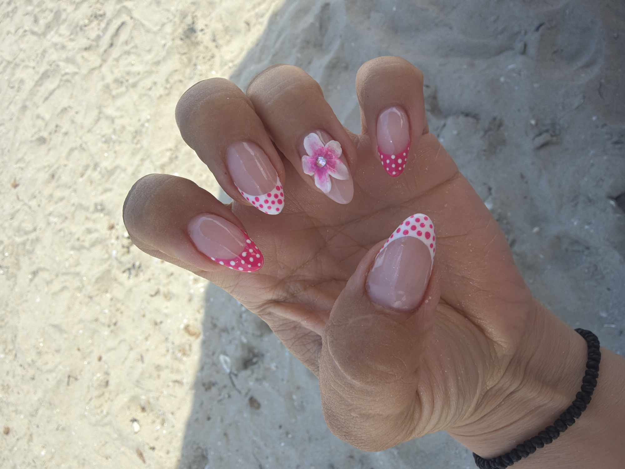 A close-up, eye-level, medium shot shows a woman's left hand with elaborate French manicured nails resting on a sandy surface. The nails are long and almond-shaped, with a pale pink base and a white tip. The tips are decorated with small, bright pink polka dots. One nail, on the ring finger, features a small, three-dimensional white and pink flower with a clear rhinestone in the center. The woman is wearing a thin, black woven bracelet on her wrist. The background is a sandy beach, with shadows cast from an unseen object. The lighting is bright and natural, suggesting an outdoor daytime setting.