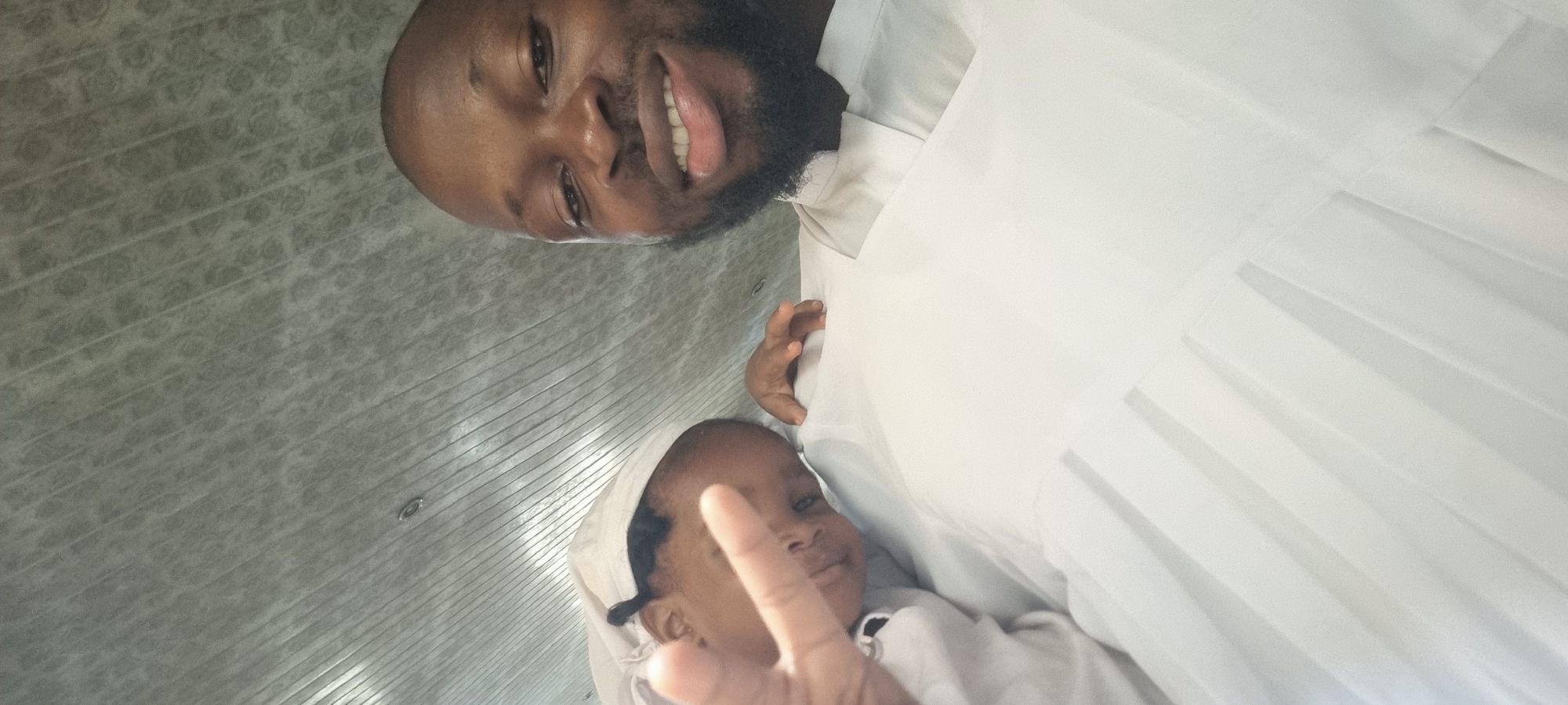 A man with a beard is smiling at the camera, holding a baby. The baby is making a "peace" sign with its fingers. Both are wearing white clothing. The setting appears to be indoors, possibly a home in Lagos, Nigeria. The wall in the background has a textured pattern.