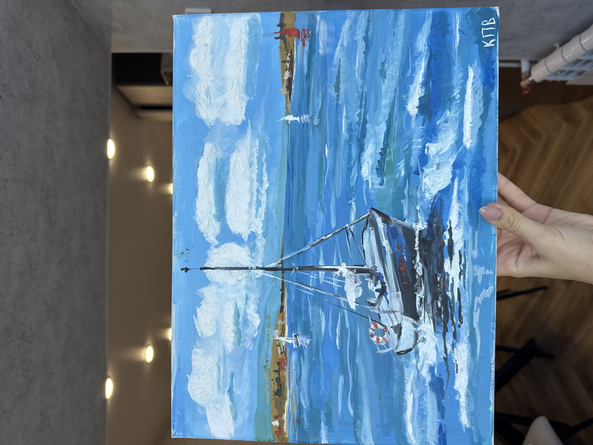 The image captures a person's hand holding up a vibrant, vertically oriented painting of a maritime scene. The painting, an acrylic or oil on canvas, depicts a sailboat on a body of water under a clear blue sky with fluffy white clouds.
The central subject of the artwork is a sailboat with a dark hull, a prominent mast, and a white lifebuoy attached to its side, positioned in the lower right section of the canvas. The water is rendered with dynamic, visible brushstrokes in various shades of blue and white, suggesting movement and reflections of light. In the upper portion, a bright blue sky is filled with textured white clouds. A distant coastline or landmass with indistinct structures, possibly lighthouses, can be seen along the horizon line. A smaller boat is also faintly visible in the distance.
The painting style is impressionistic, characterized by noticeable brushwork that adds texture and movement, particularly to the water and clouds. In the upper right corner of the painting, the Cyrillic letters "КПВ" are painted in white.
The hand holding the painting is visible on the right, featuring manicured nails with a light polish. The background beyond the painting and hand includes a grey textured wall on the left, several warm circular lights illuminating a surface in the mid-ground, and glimpses of a wooden floor and a white radiator in the upper and lower right corners, indicating an indoor setting. The painted scene depicts a bright, clear day.