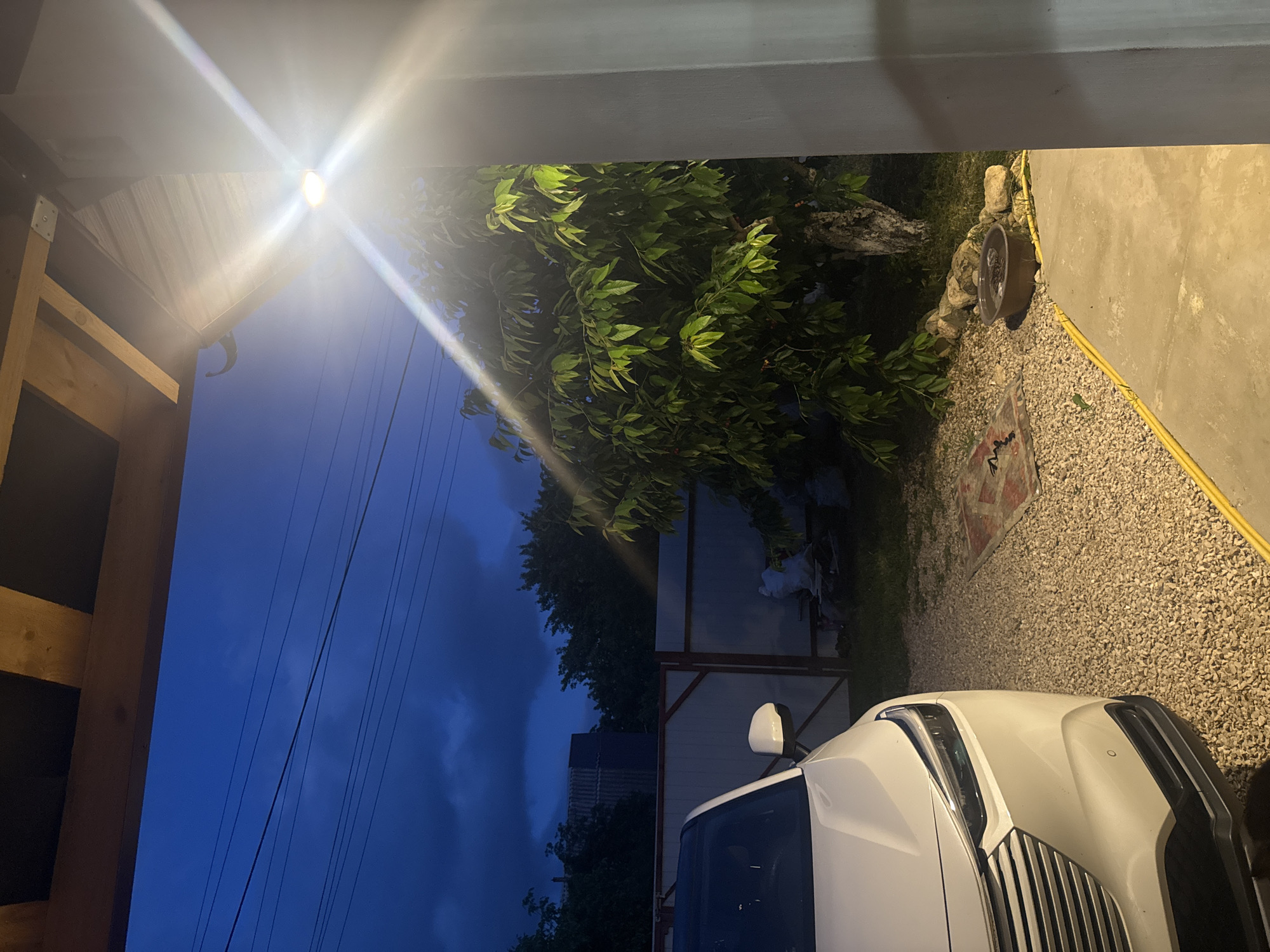 The image depicts a nighttime scene in what appears to be a residential yard in Добровское сельское поселение, Ukraine. In the foreground, the front section of a white car is visible on the left. The car is parked on a gravel surface. To the right, a concrete patio leads towards the viewer, bordered by a yellow hose. Beyond the car, a fence runs across the middle ground, with a large tree to its right. The tree is illuminated by a bright light source, possibly an outdoor lamp, which creates strong lens flares and highlights. The background shows a dark, cloudy sky with visible power lines. In the far background, a building with a corrugated roof is discernible. Several objects are scattered in the yard, including a rug with a pattern, a circular metal basin filled with water and rocks, and a white bag. The overall atmosphere is dim, with the artificial light creating a stark contrast with the natural darkness.