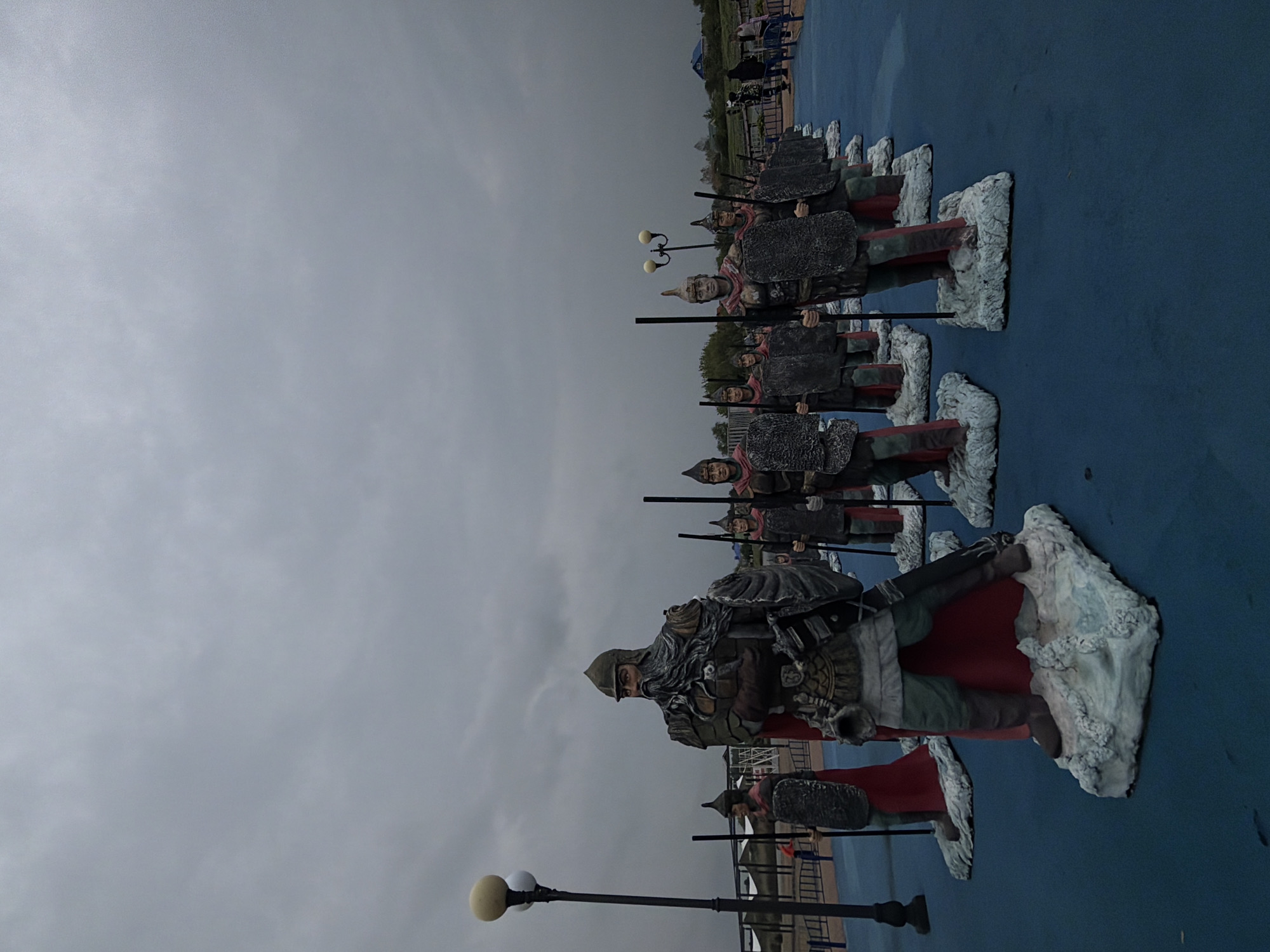 The image shows a row of statues of ancient warriors on a blue surface. The statues are standing in formation, each holding a spear. The statues are standing on what looks like white clouds. The background is a gray sky. The location is unknown.