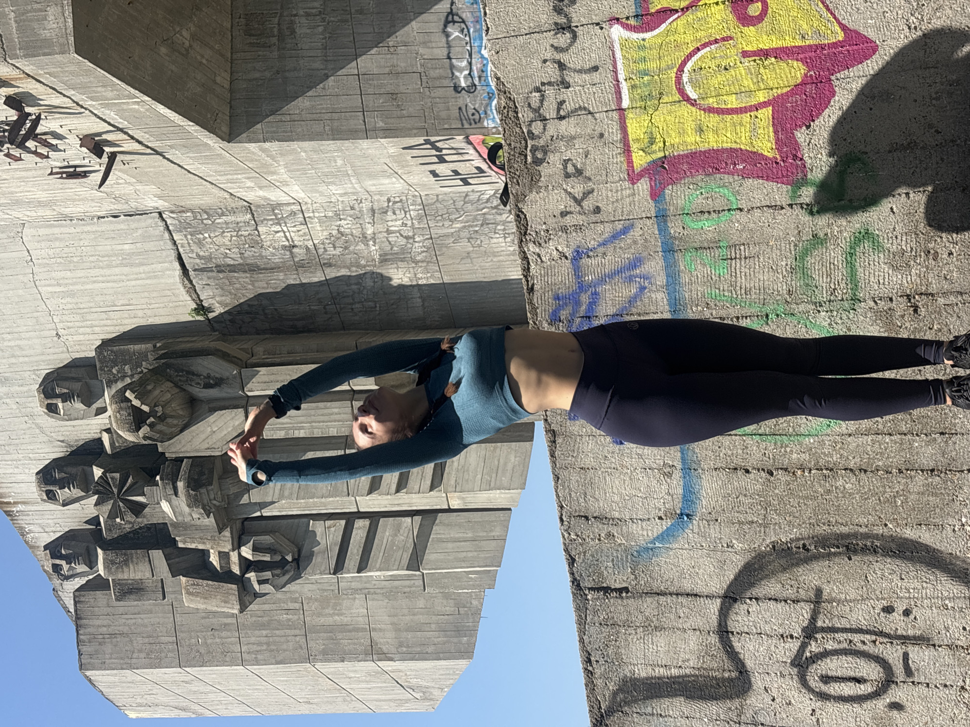 A full-length vertical shot captures a young woman in an upward stretch pose against the concrete backdrop of the Monument to the Bulgarian-Soviet Friendship in Varna, Bulgaria.
The woman stands centered in the lower half of the frame, facing slightly left with her head tilted back and eyes looking upward. Her arms are raised above her head, hands clasped together with fingers intertwined. She wears a long-sleeved, teal-colored athletic crop top that exposes her midriff, and dark navy or black athletic leggings paired with dark sneakers. Her hair, styled in two braids, falls over her shoulders.
Behind the woman and dominating the upper left and center-right of the image is the massive, brutalist concrete structure of the monument. The monument features several stylized, abstracted human figures sculpted into its facade, with two prominent busts visible in the upper left section. The concrete exhibits significant weathering, discolored patches, and visible cracks. On the right side of the monument, above a large archway and slightly hidden by shadow, are several abstract, metallic, rebar-like elements protruding from the concrete surface.
In the immediate foreground, running horizontally across the lower third of the image, is a concrete wall or barrier, heavily defaced with various graffiti. Notable graffiti includes large black outline letters, a bold yellow and pink bubble letter "G", a light blue curved line, and the block letters "HE HA" spray-painted in black on a section of the monument's lower wall behind the foreground barrier. The ground is not visible, with the wall forming the bottom edge of the image.
The scene is brightly lit by direct sunlight from the upper right, casting sharp shadows on the monument's features, the wall, and a distinct shadow of the woman's head and upper body on the graffiti-covered wall to her right. The sky, visible in the upper left, is a clear, bright blue.
