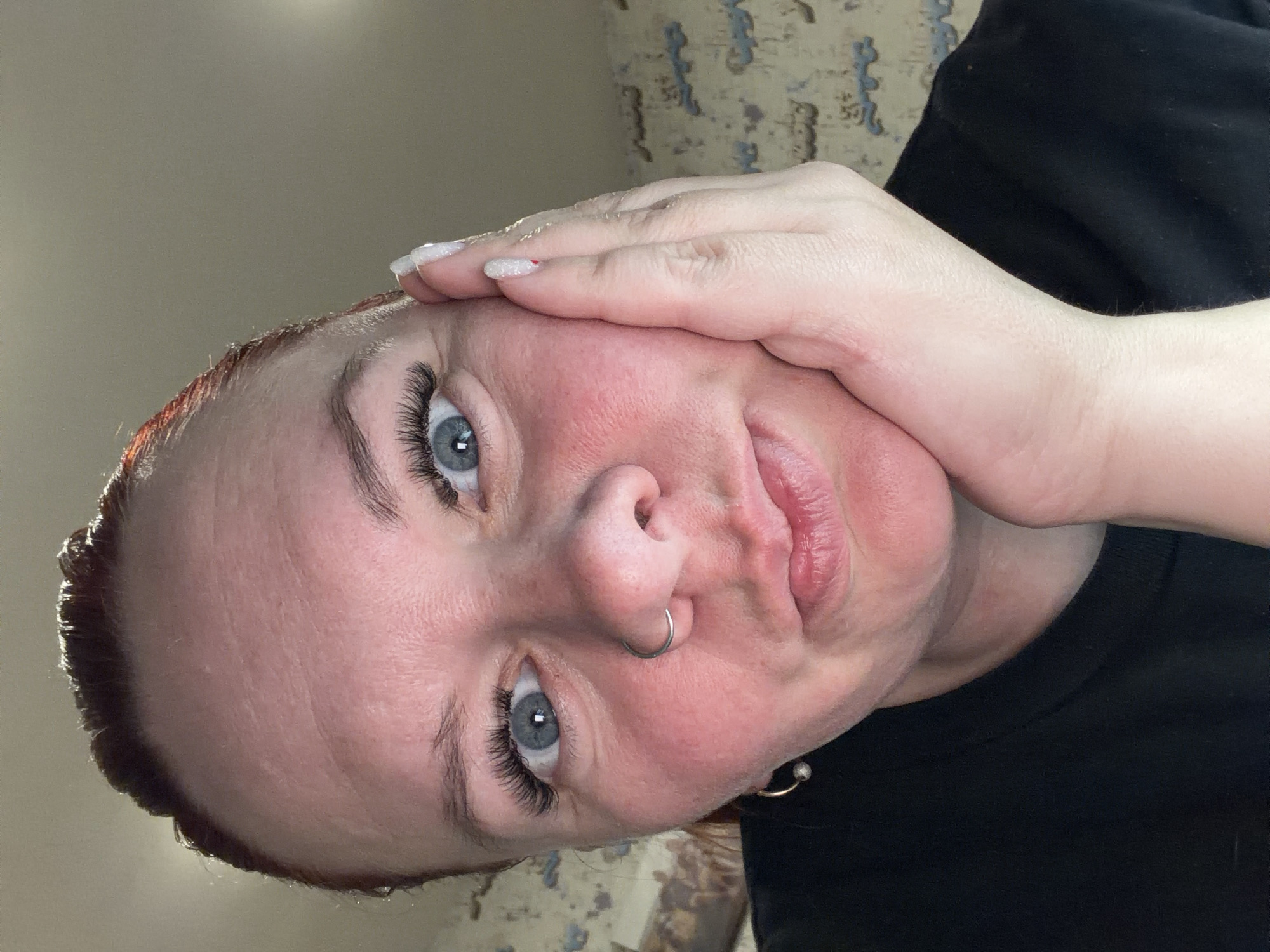 A close-up, rotated selfie features a woman with fair skin, light blue eyes enhanced with eyelash extensions, and reddish-brown hair. She has a nose ring and an earring visible on her left side. Her nails are painted white with silver glitter. Her face is slightly flushed. Her left hand is resting on her forehead, and she's wearing a black garment. The background appears to be a patterned wall and a plain light-colored wall. There is no verifiable information to confirm this image was taken in the city of Chaikovsky, Russia.