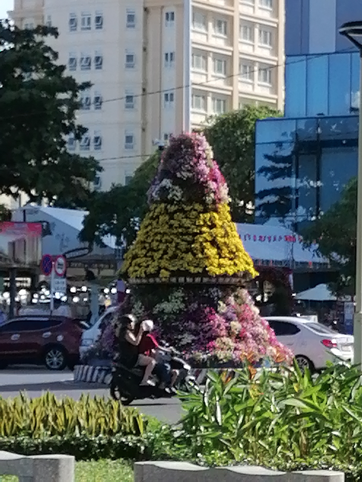 The image depicts a street scene in Nha Trang Ward, Vietnam. A large, tiered floral display dominates the foreground. The display features multiple colors of flowers, including pink, white, yellow, and purple, arranged in a pyramid shape. Behind the floral display, there is traffic on a street, including cars and a scooter with two people riding it. The background consists of buildings, including a tall, light-colored building with numerous windows and a blue glass-fronted building. Green foliage is present in the foreground and scattered throughout the scene, providing a natural element within the urban environment. Traffic signs are visible in the background, indicating the presence of a busy street. The overall lighting suggests it is daytime.