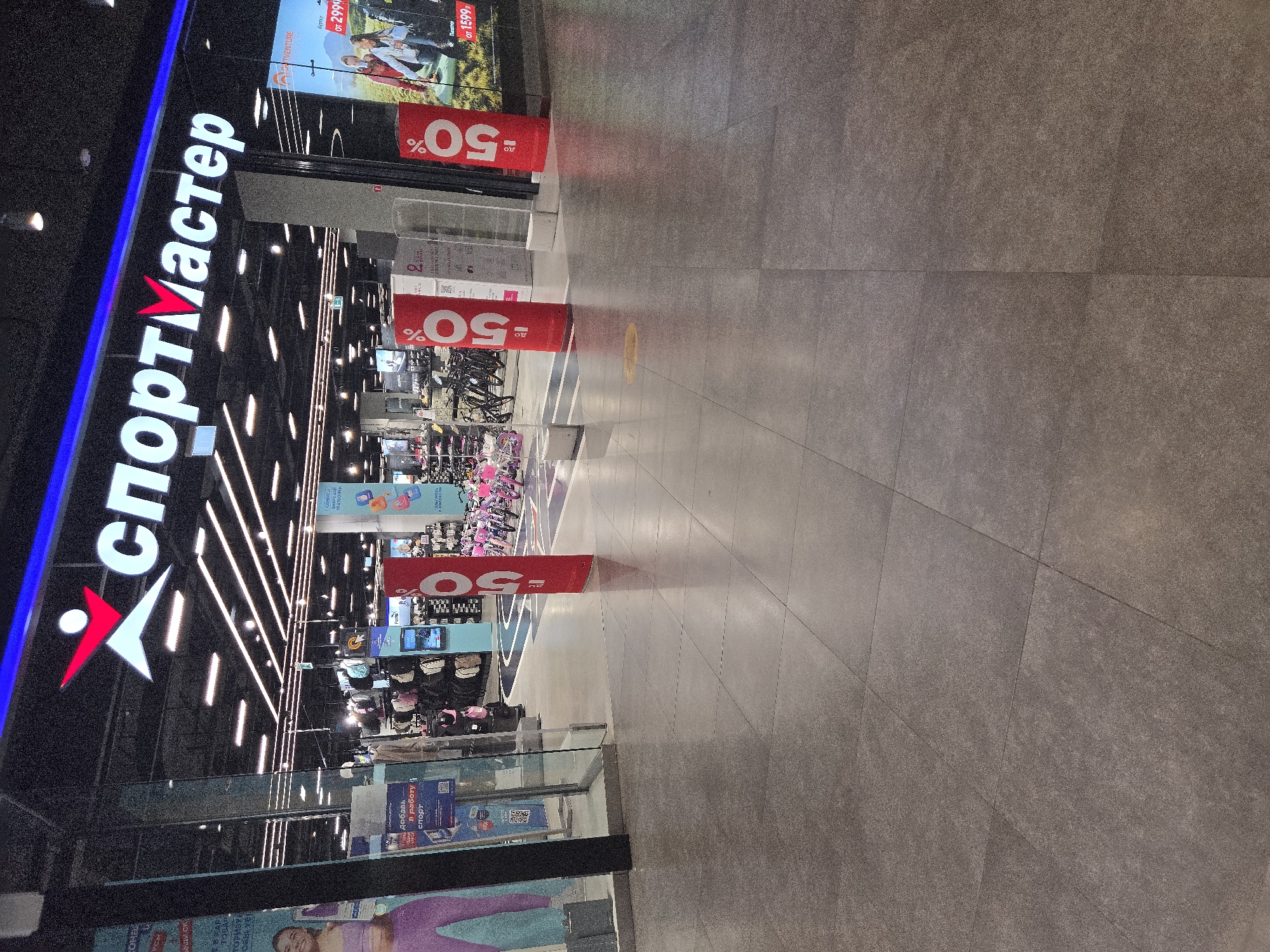 This is a full-frame, eye-level, medium shot of the entrance to a Sportmaster store, a Russian sports and outdoor goods retailer. The store is located in Moscow, Russia. The storefront features a large illuminated sign that reads "СПОРТМАСТЕР" (SPORTMASTER) in white letters with a red stylized person logo above. The entrance is wide, with glass doors and a glass-paneled exterior.
Inside the store, rows of bicycles, primarily children's models in bright colors, are visible. Merchandise, including backpacks and other sports equipment, is displayed on shelves and racks. Two tall, cylindrical banners with "до -50%" (up to -50%) printed in white on a red background are placed outside the entrance, suggesting a sale.
A poster on the right side of the entrance displays images of people engaged in outdoor activities and shows pricing information: "от 2999р." (from 2999 rubles) and "от 1599р." (from 1599 rubles). To the left, a display includes a QR code and text, likely an advertisement or information panel.
The floor in front of the store is tiled with large, square gray tiles. The lighting is a mix of overhead fluorescent lights and accent lighting from the store's signage. There are no people clearly visible in the immediate foreground or entering/exiting the store. The overall impression is that of a retail environment.