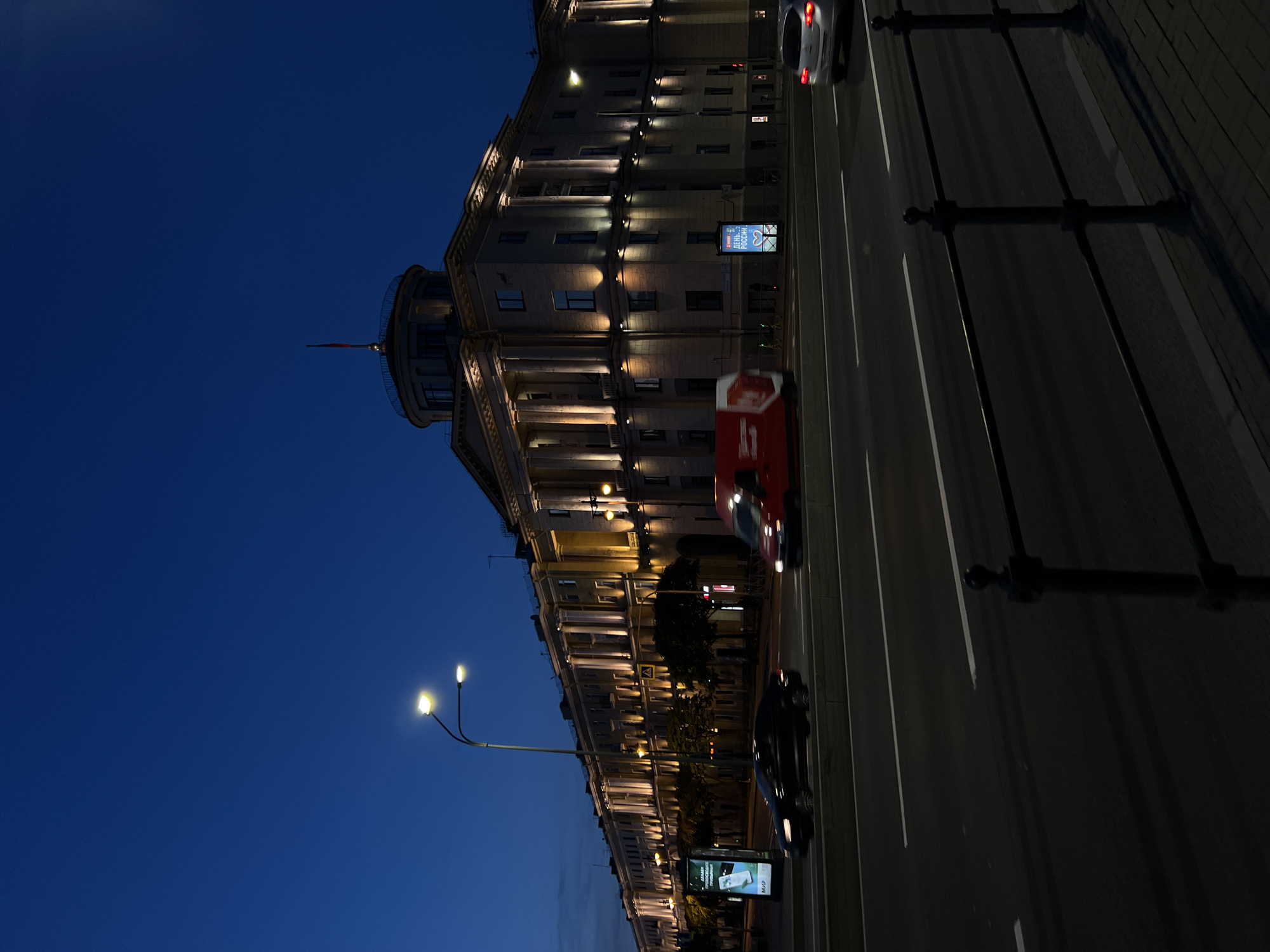 An eye-level, medium shot captures a grand building in a city at dusk. The building, illuminated by exterior lights, features classical architectural elements such as columns and a domed structure topped with a flag. A street with multiple lanes runs in front of the building, with cars in motion, indicating traffic. Streetlights and advertising billboards are also visible. The sky is a deep blue, transitioning from daylight to night. The overall scene depicts an urban environment at twilight, with a focus on the illuminated architecture and the flow of city life.