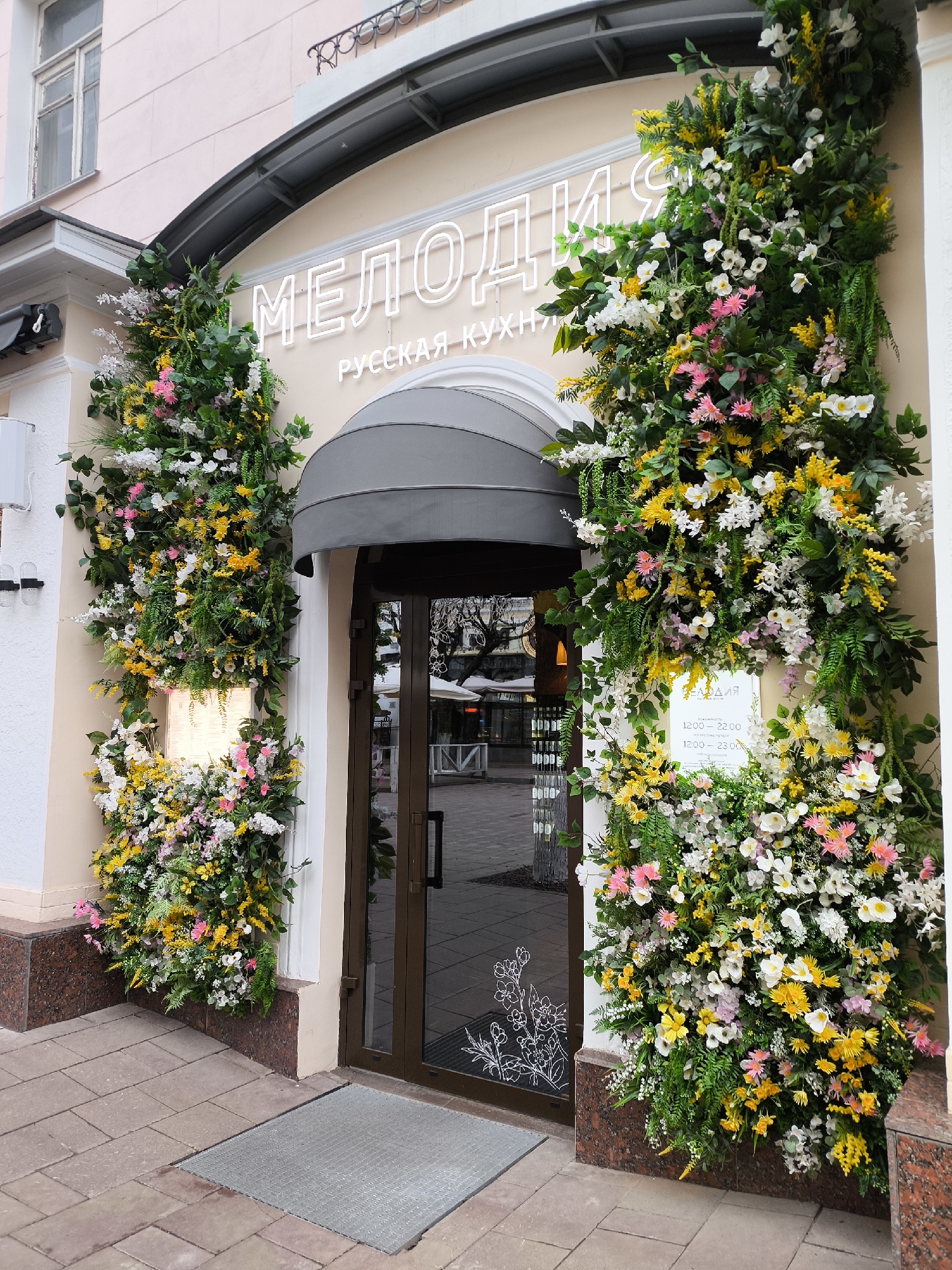 The image shows the entrance of a restaurant named "MELODIYA" with the words "RUSSIAN CUISINE" written below. The entrance is framed by floral arrangements composed of white, yellow, and pink flowers intertwined with green foliage. A grey awning is located above the glass door. On the door, a small white flower outline is visible. A sign on the right side of the entrance displays the opening hours of the restaurant. The exterior facade of the restaurant is painted in light shades with marble base. Outside, the sidewalk is paved with concrete blocks, and a metal grate is set into the pavement in front of the door. The location is in a city of Unknown, Unknown.