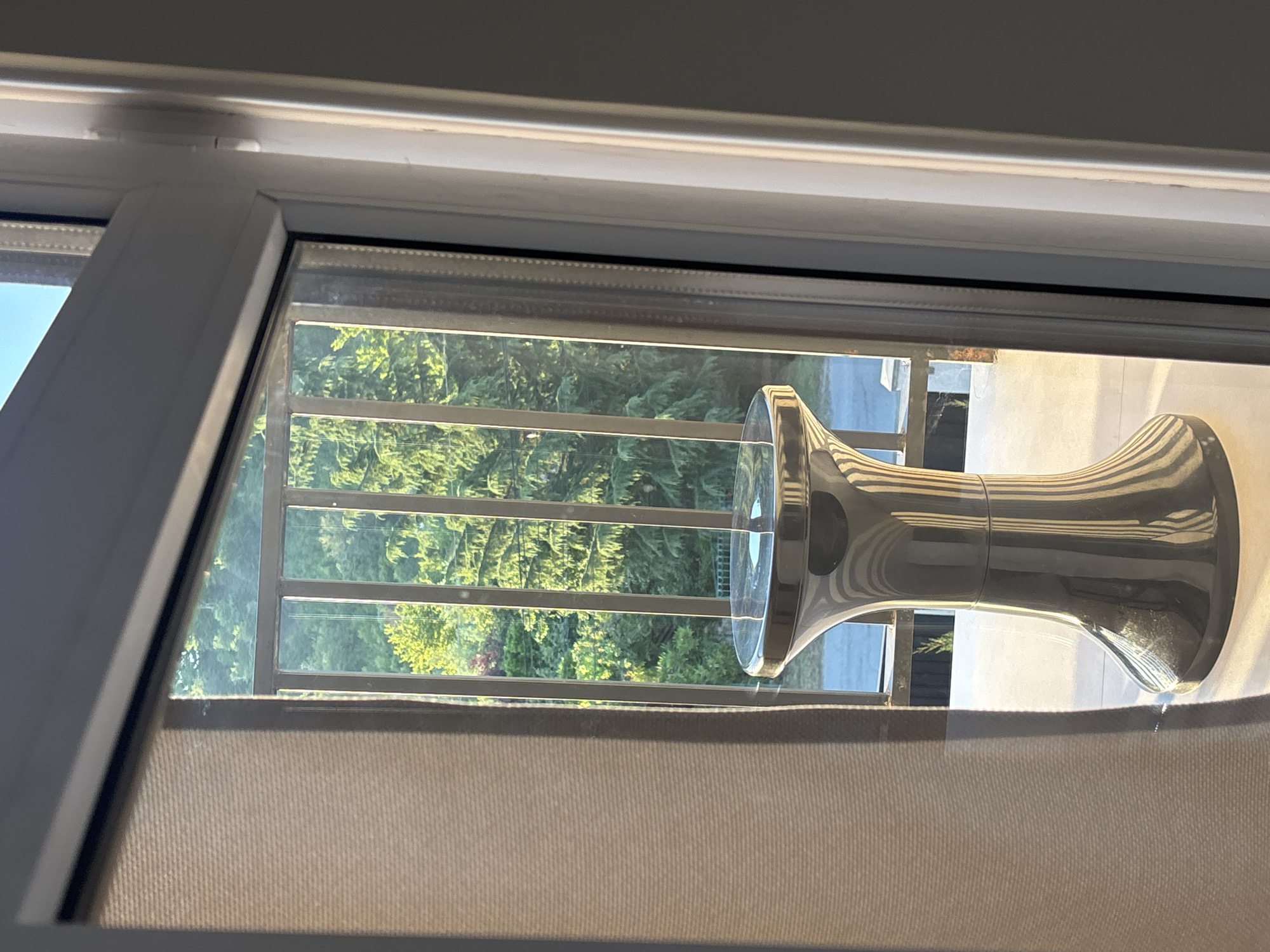 The image shows a silver, hourglass-shaped stool placed on a light-colored floor, viewed through a window from inside a building. The stool is highly reflective, showing vertical stripes of light and shadow across its surface. Behind the stool, a metal railing is visible, beyond which a dense forest of green trees extends upwards towards a bright blue sky. A beige curtain hangs on the left side of the frame, partially obscuring the view. The scene suggests an outdoor setting, possibly a balcony or patio, with a lush, natural environment visible. The lighting indicates it is daytime, with sunlight casting sharp shadows. The provided context of Ldzaa, Abkhazia, places the scene in a specific geographic location known for its scenic beauty and natural landscapes.