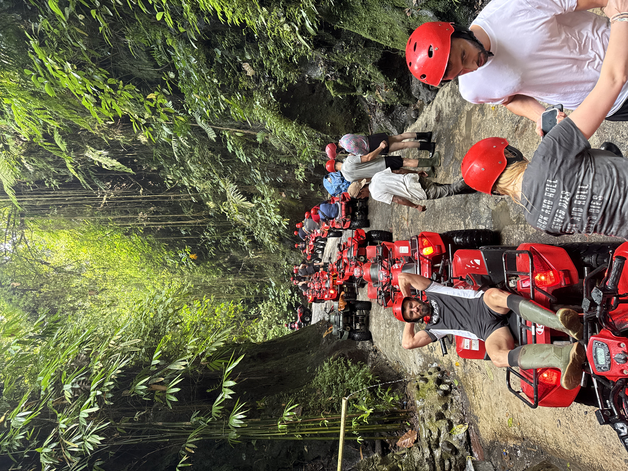 A group of approximately twenty individuals on all-terrain vehicles (ATVs) is halted on a narrow, wet, and muddy path within a deep, verdant gorge in Payangan, Indonesia. The path is flanked on both sides by dense, tropical rainforest vegetation, featuring tall bamboo stalks on the left and a thick wall of ferns, moss, and climbing vines on the right.
In the immediate foreground, a man with a beard and eyeglasses, wearing a black and white athletic shirt, black shorts, and tall olive-green rubber boots, reclines on a red ATV with his hands clasped behind his head. He is wearing a red helmet. To his right, another man, also with a beard and wearing a white t-shirt, black shorts, and a red helmet, holds a smartphone, which a woman in a grey t-shirt with "ROCK AND ROLL NEVER DIES" printed on the back and a red helmet is looking at.
A long line of mostly red ATVs, interspersed with one dark green ATV, extends into the background, where more riders, all wearing red helmets, are visible. Many individuals wear similar green or black rubber boots. The ground is wet, showing puddles and signs of recent water flow. On the left side of the path, a natural water spout, possibly a bamboo pipe, directs a stream of water into a small channel on the ground. The lush canopy overhead creates dappled lighting throughout the scene. The group appears to be on a recreational ATV tour, currently paused amidst the dense jungle environment.