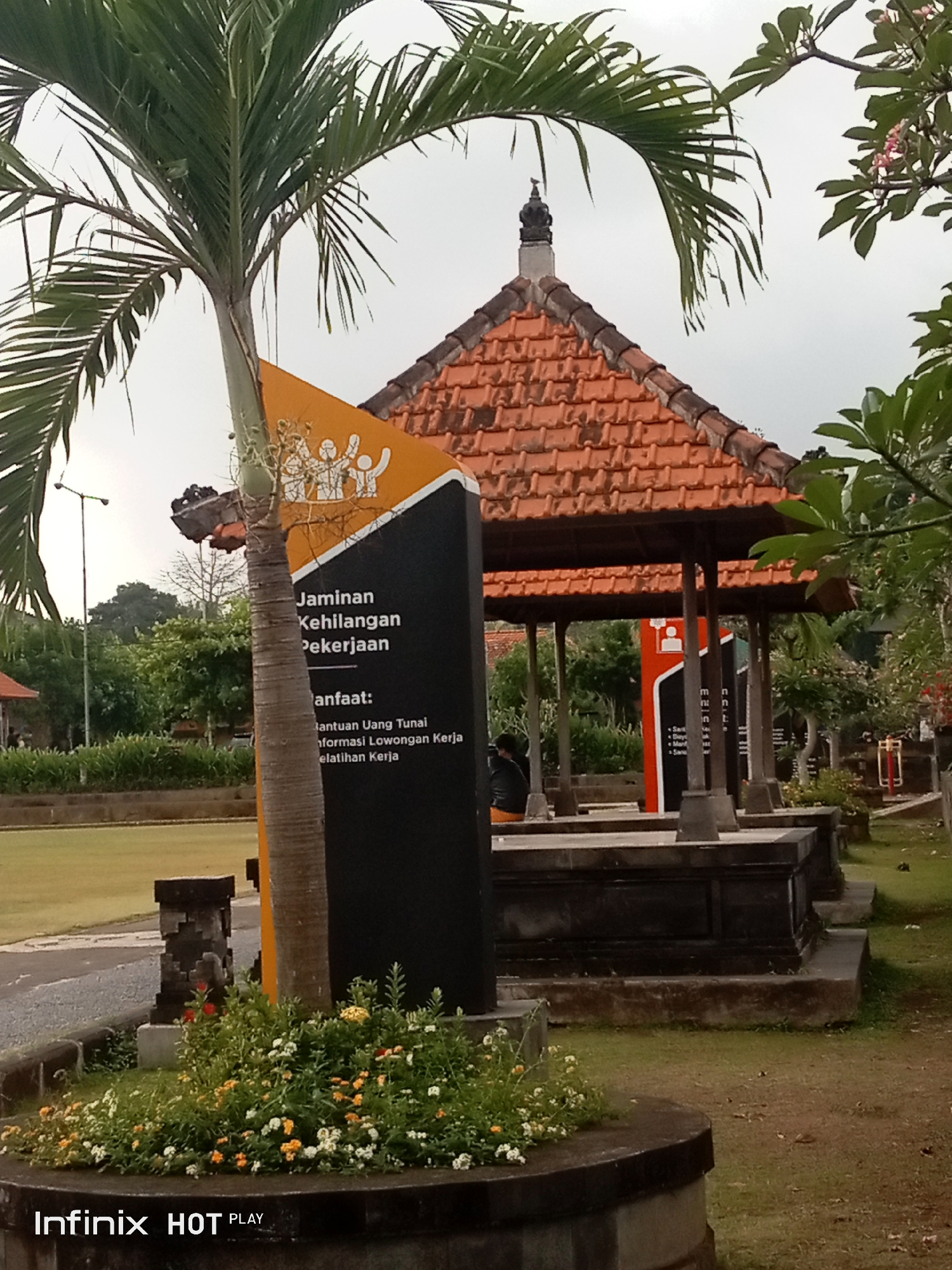 The image depicts an outdoor scene with traditional Balinese architecture. In the foreground, a palm tree with lush green fronds stands to the left, partially obscuring a black sign with yellow text and orange graphics. The sign contains Indonesian text, including "Jaminan Kehilangan Pekerjaan" which translates to "Unemployment Insurance" and "Manfaat" meaning "Benefits." Beneath "Manfaat," the text lists "Bantuan Uang Tunai" (Cash Assistance), "Informasi Lowongan Kerja" (Job Vacancy Information), and "Pelatihan Kerja" (Job Training).
Behind the sign is a traditional Balinese pavilion with a terracotta-tiled roof, supported by wooden posts. The pavilion is elevated on a stone base. In the mid-ground, there are stone structures that appear to be part of a monument or seating area. Several people are visible in the distance, some sitting within the pavilion and others walking in the park-like setting.
The environment is green and well-maintained, with a grassy lawn and manicured bushes with small white and orange flowers in the foreground. In the background, there are more trees and some buildings. The sky is overcast. The overall impression is that of a public space or park in a tropical location, likely in Blahbatuh, Indonesia, given the architectural style. A watermark at the bottom of the image reads "Infinix HOT PLAY."