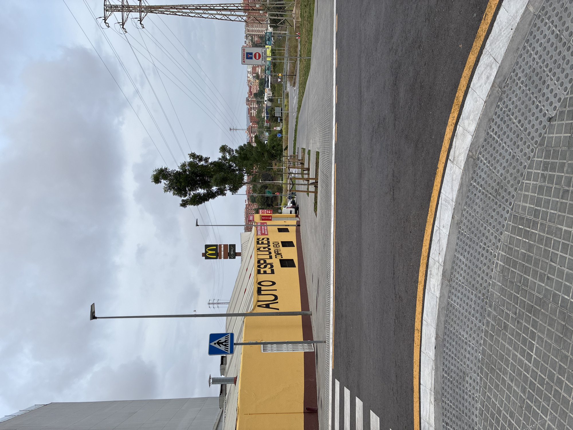 The image captures an outdoor urban street scene in Esplugues de Llobregat, Spain, under an overcast sky during daytime. The perspective is tilted, showing a road, sidewalk, and various structures.
In the immediate foreground, a dark asphalt road with a prominent yellow line and a white line defining its edge curves gently to the right. Adjacent to the road is a grey tiled sidewalk, featuring both square and circular patterned paving stones. A white zebra-striped pedestrian crossing is partially visible in the lower left.
The mid-ground is dominated by a long, low yellow building with black text painted on its side, reading "AUTO ESPLUGUES" and "COMPRA VENTA" (Buy Sell). A blue square sign with a white pedestrian crossing symbol is mounted on a pole directly in front of this building. Above the building, a yellow McDonald's 'M' logo is visible on a tall signpost. Further down the street, a red and white traffic sign displays "ATENCIO" (Attention), a "No Entry" symbol ("SENTIT PROHIBIT"), and "EXCEPTE ACCES MAU" (Except Maintenance Access). A white van is parked near the building's entrance, which has a partially visible red sign marked "ENTRADA" (Entrance).
To the right of the yellow building, a paved pathway lines a grassy strip with small, sparsely leafed trees. In the background, numerous residential buildings with varied colors can be seen on a distant hillside. Overhead, a complex network of power lines stretches across the cloudy grey sky, supported by a large metal electricity pylon on the left. The overall impression is one of a functional commercial/industrial area bordering a residential zone.