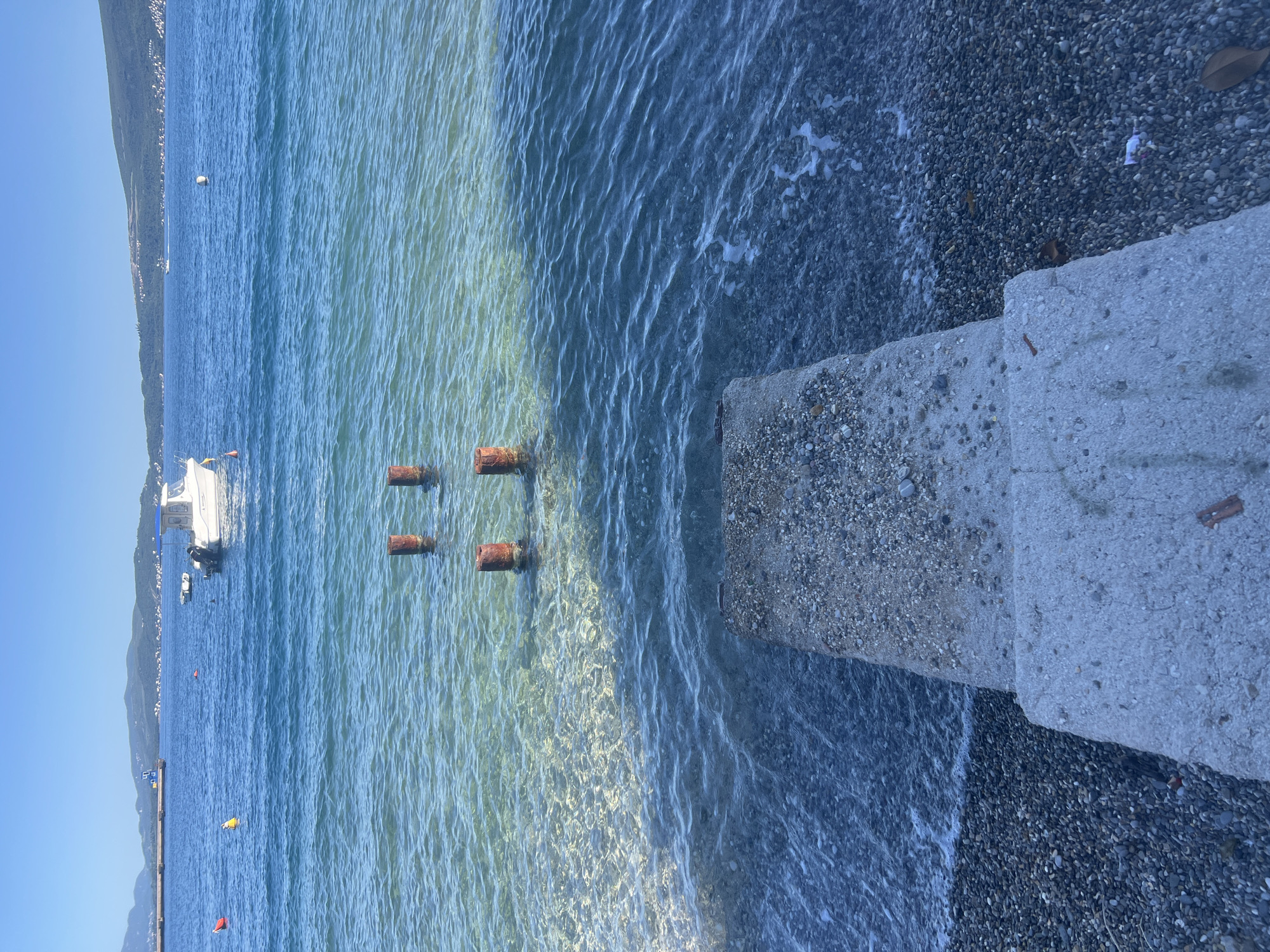 The image depicts a waterfront scene in Bijela, Montenegro, featuring a concrete pier extending into the water. The water is clear, with some turbidity, and several rusty cylinders are partially submerged. A beach composed of small, dark pebbles borders the water's edge. In the background, a boat is docked, with mountains visible along the coastline. The sky is clear and blue.
