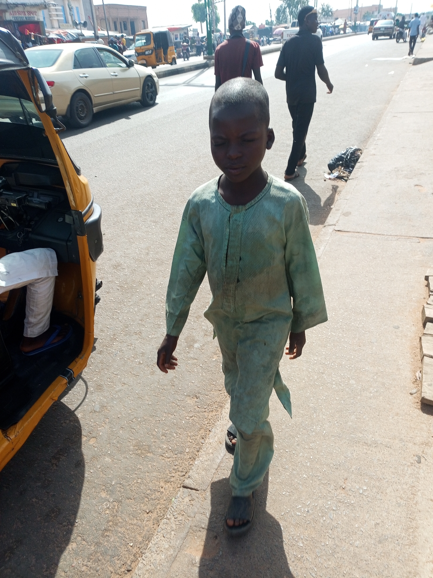 The image depicts a bustling street scene in Tempure, Nigeria, under bright, clear sunlight. The central focus is a young boy, appearing to be between 5 and 8 years old, with dark skin and short dark hair. He is walking towards the viewer on a concrete sidewalk, wearing a light green, somewhat faded and possibly soiled, traditional-style long-sleeved tunic and matching trousers, paired with dark sandals. His eyes are downcast, giving him a neutral or somber expression.
To the immediate left, the front section of a yellow motorized tricycle (keke napep) is visible, with a person's legs, clad in white trousers and blue and brown sandals, seen inside the driver's compartment. The engine area of the tricycle appears to be open.
The background shows a paved road with vehicular and pedestrian traffic. A golden sedan car is driving away from the camera, and another yellow tricycle can be seen further down the street. Several individuals are walking in the mid-ground; two men are particularly noticeable walking away from the camera, one in a dark red shirt and a patterned head covering, and another in a black shirt with a white headband. On the right side of the sidewalk, a small heap of trash, including a black plastic bag, is present.
The strong, bright sunlight creates distinct, long shadows on the ground, suggesting it is midday or early afternoon. In the background, a red sign on a building reads "ADE SULA SHOPT," and "TO" is partially visible on a white vehicle further down the street. The scene conveys an everyday moment in an urban setting.