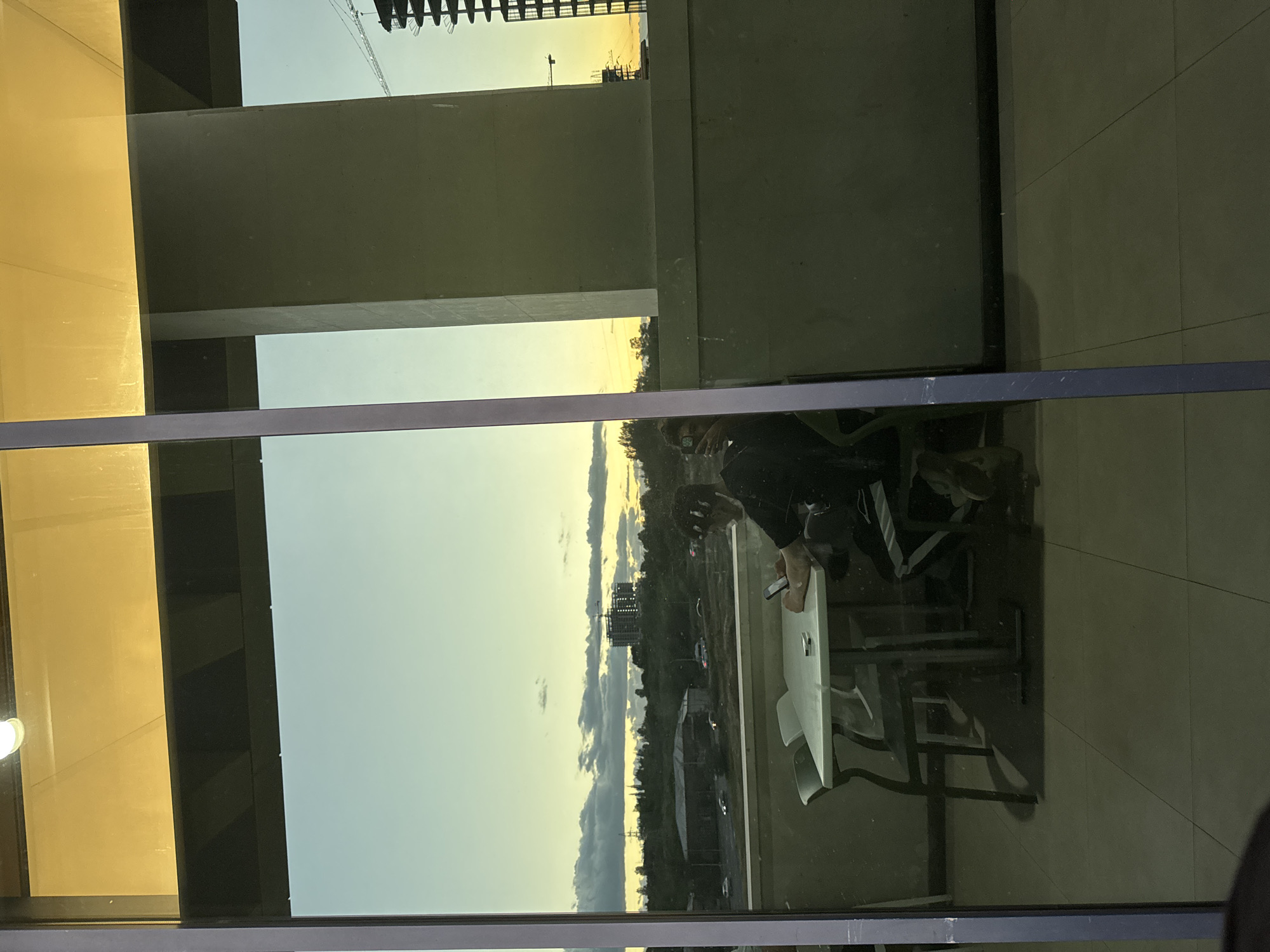 The image captures a view through a large glass window or partition, simultaneously revealing interior reflections and the exterior landscape. The scene is oriented vertically in the provided image, but when viewed upright, it shows an indoor perspective looking outwards.
In the foreground, reflections of an indoor setting are prominent. A person is seated at a table with a light-colored top and dark base, holding a mobile phone in their hand. Parts of a chair are also reflected.
Beyond these reflections, the exterior view depicts a sky transitioning from a light blue at the top to a warm golden and orange hue near the horizon, strongly suggesting either sunrise or sunset. Scattered clouds are visible against this gradient sky. The landscape beneath the sky includes distant urban structures, with a prominent crane indicating ongoing construction in one area. Other buildings and a wider cityscape or landscape are visible in the distance, implying an urban or peri-urban environment. A dark, thick horizontal beam or frame element spans across the middle of the window.
The time of day is a notable detail, characterized by the warm, atmospheric lighting. The setting appears to be an urban location, potentially in Yerevan, Armenia, as indicated by the context. No discernible text is visible in the image.