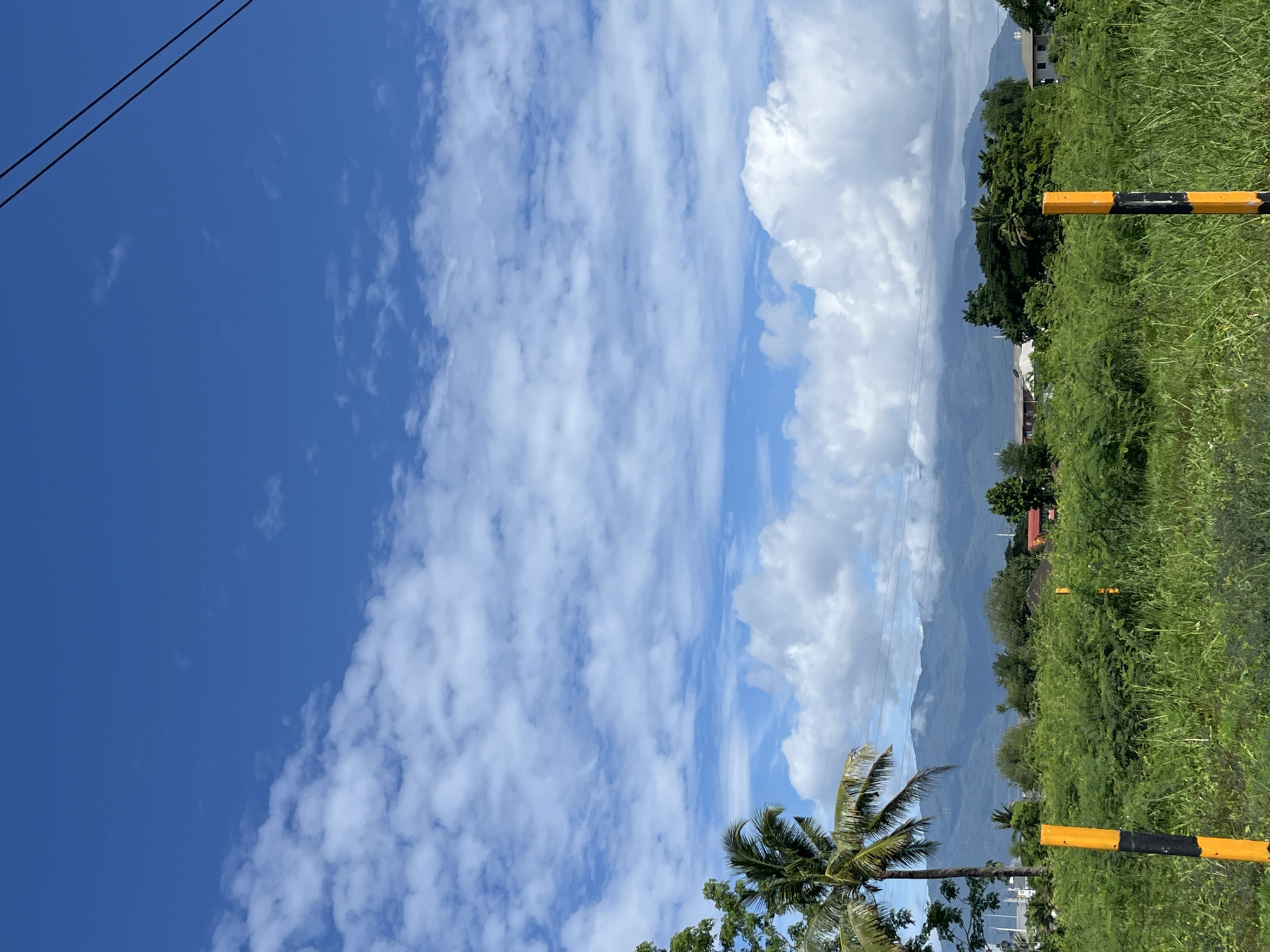The image shows a landscape scene from the city of Ban Kanchanok 23, Thailand. The sky is a vibrant blue with scattered white clouds. In the foreground, there is a field of lush green vegetation, with two yellow and black striped posts visible. A tall palm tree stands to the left, and beyond the vegetation, there are several houses with red roofs and other buildings nestled amongst trees. In the distance, a range of mountains is visible, partially obscured by clouds. Power lines stretch across the upper right portion of the frame.