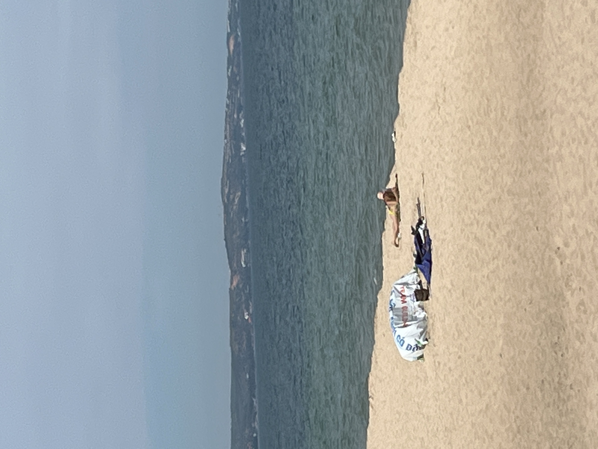 This is a wide shot of a beach in Mũi Né, Vietnam, on a hazy day. The water is a deep blue, and the sky is a pale, hazy blue. In the foreground, there is a sandy beach with a few items. A woman in a yellow bikini is lying on her stomach near the water's edge, facing away from the camera. Further to the left, there is a large white and blue patterned bundle, possibly a beach umbrella or tent, on the sand. Next to the bundle, there are some dark blue items of clothing or towels. In the background, a distant, undulating coastline with sparse buildings is visible across the water.