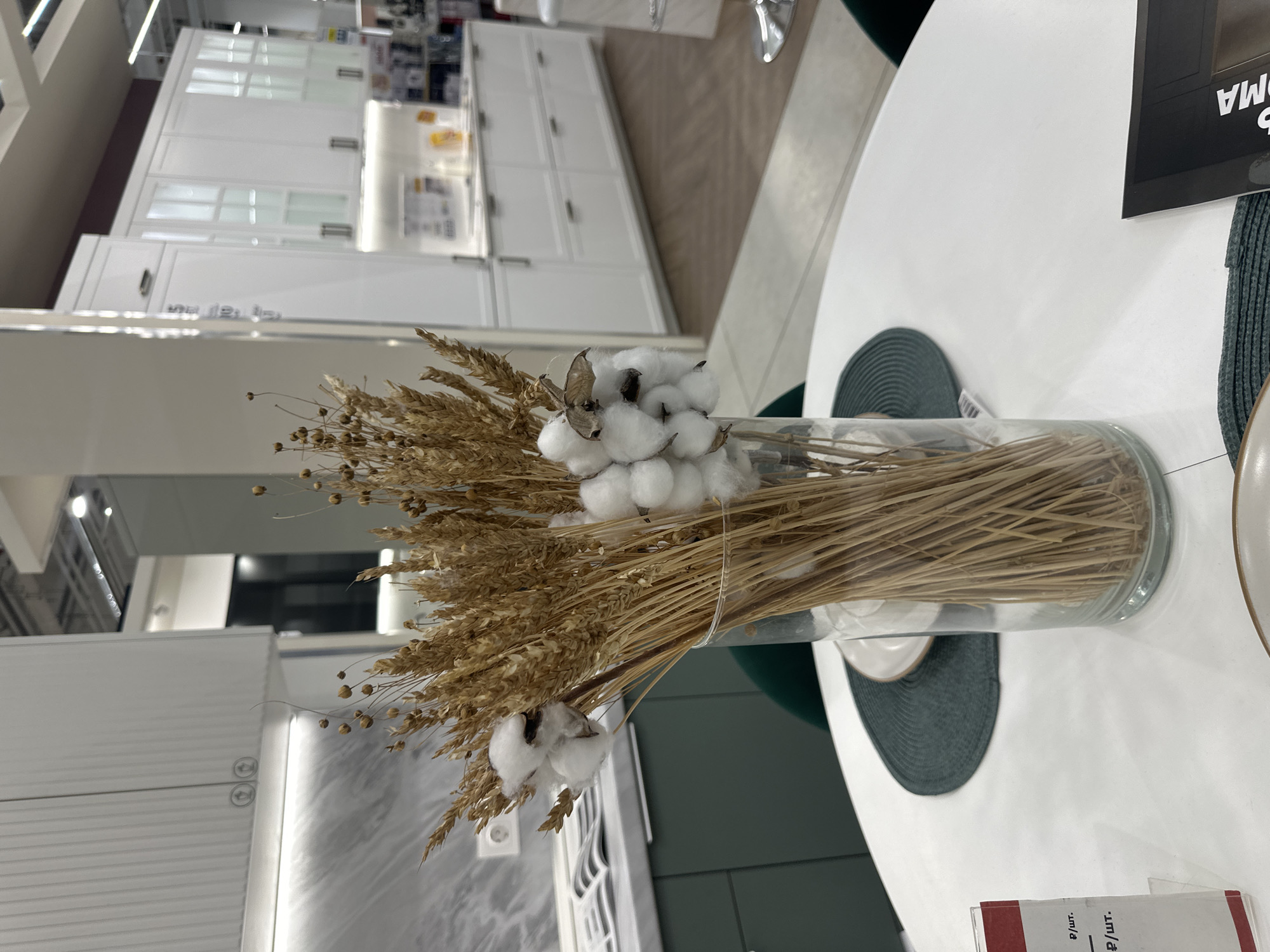 The image shows a still life of a bouquet in a cylindrical glass vase, arranged on a white circular table. The bouquet comprises dried wheat stalks and tufts of cotton. Two dark-green, circular placemats lie under the vase. A white plate is partially visible on the right side of the table. In the background, there are white kitchen cabinets and a partial view of what appears to be a retail setting or showroom. A card with text in Russian is visible at the bottom of the frame. Based on the product context and store design, this scene may be set in an IKEA store in Moscow, Russia.
