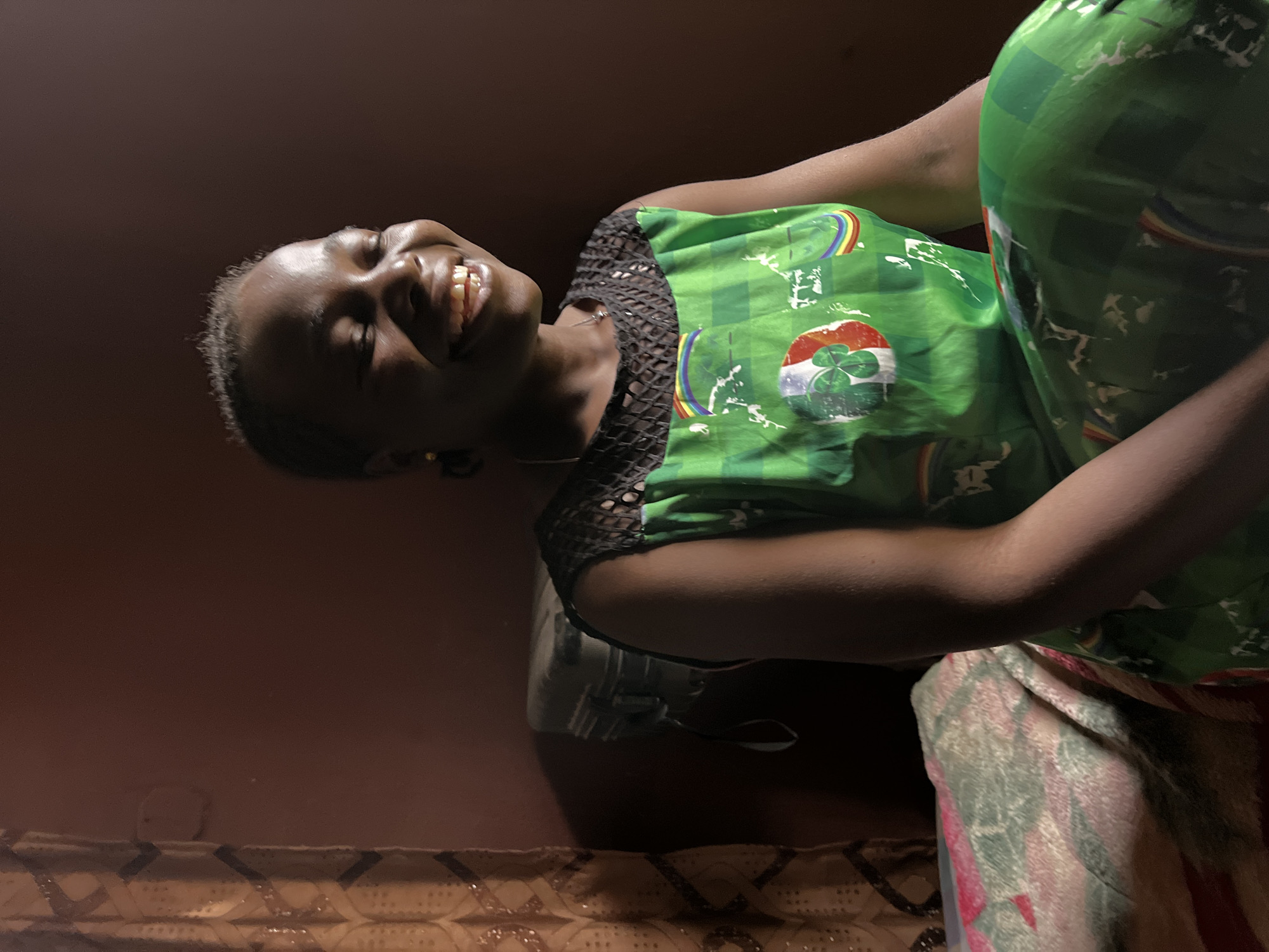 The original image, captured in Jos, Nigeria, features a young woman with dark skin and short dark hair, shown in a low-light indoor setting. She is positioned diagonally, looking upwards and to the left with a wide, open-mouthed smile that reveals her teeth, conveying genuine happiness and joy.
She wears a vibrant green top or dress adorned with multiple graphic elements, including stylized rainbows and a prominent circular emblem. This emblem features a three-leaf shamrock overlaid on a design that resembles the Irish flag with green, white, and orange stripes. The neckline of her garment is a dark, possibly crocheted or mesh, material.
The background consists of a dark reddish-brown wall. Below her, patterned textiles are visible: one directly beneath her in shades of pink, green, and white, and another further down with an earthy, geometric pattern, likely a rug or mat on the floor. A faint, blurry reflection of an object, possibly a container, can be seen near her left arm. The intimate and cheerful atmosphere is highlighted by the subject's radiant expression in the dimly lit environment.