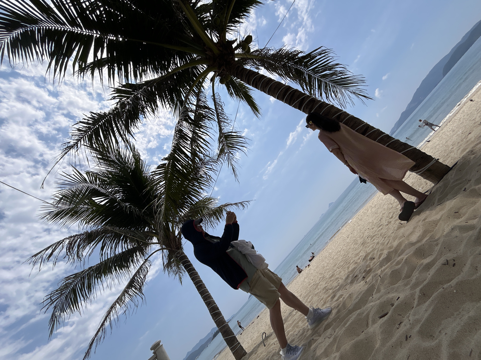 A low-angle, wide-shot photo shows a beach scene in Nha Trang, Vietnam. Two palm trees are the primary focus, one on the left and one on the right.
A man stands near the left palm tree, clad in khaki shorts, light-colored sneakers, and a dark hooded jacket. He appears to be taking a photo with his phone.
A woman stands next to the palm tree on the right, dressed in a light pink dress and sunglasses.
The sandy beach stretches out in the foreground, with the ocean visible in the background. The sky is blue with scattered clouds. There are other people on the beach in the background. Distant mountains are visible on the horizon.